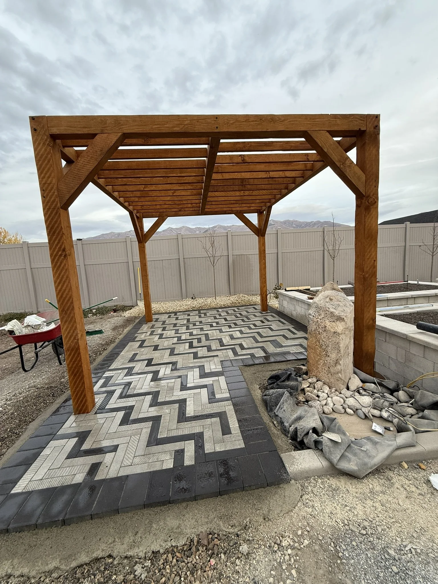 A backyard with a newly constructed wooden pergola, a patterned brick walkway, and a landscaped area with rocks and a stone feature, enclosed by a white fence.