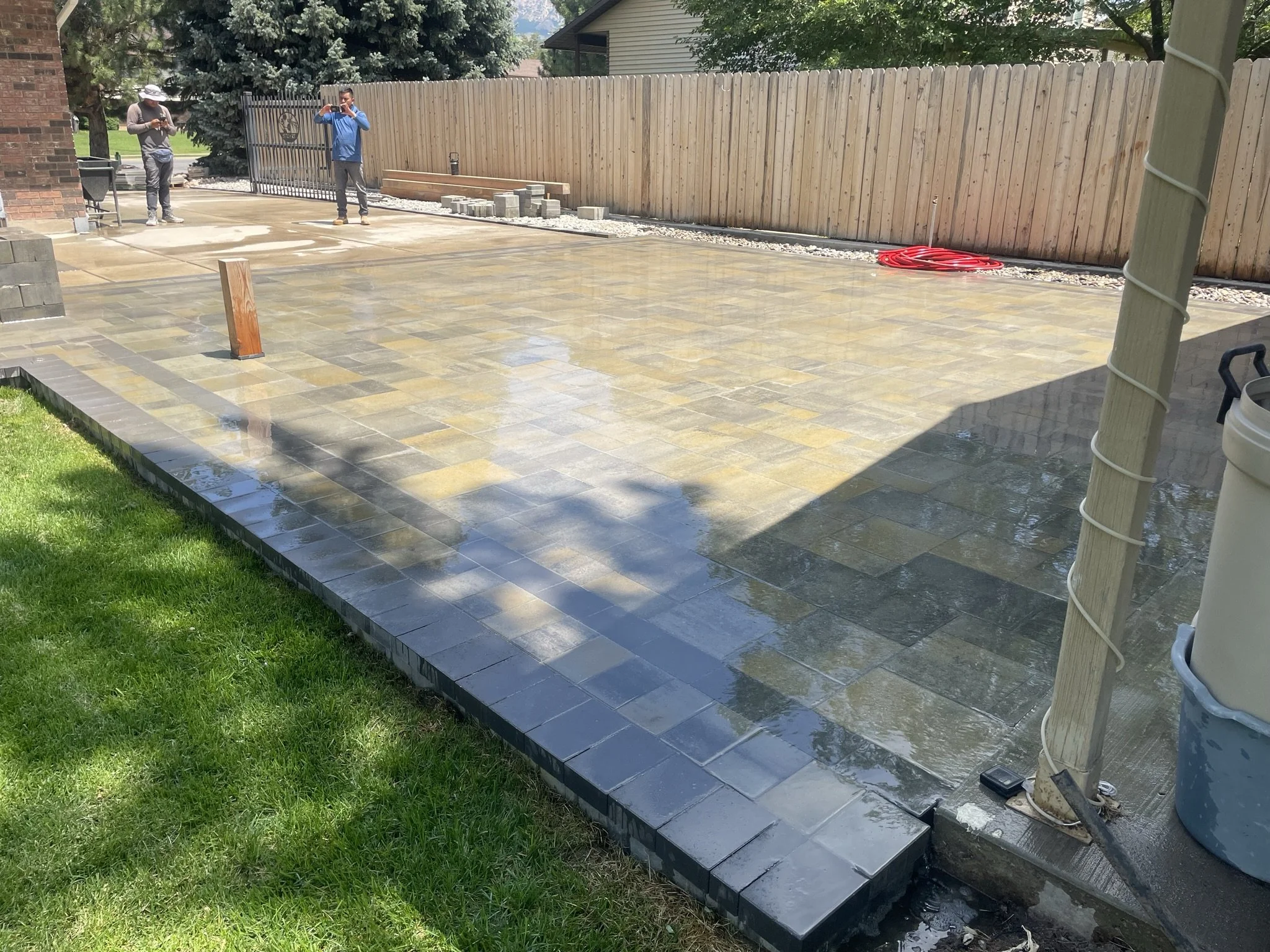 A backyard with a newly paved stone patio, some construction tools, and workers working on the project, with a wooden fence and trees in the background.