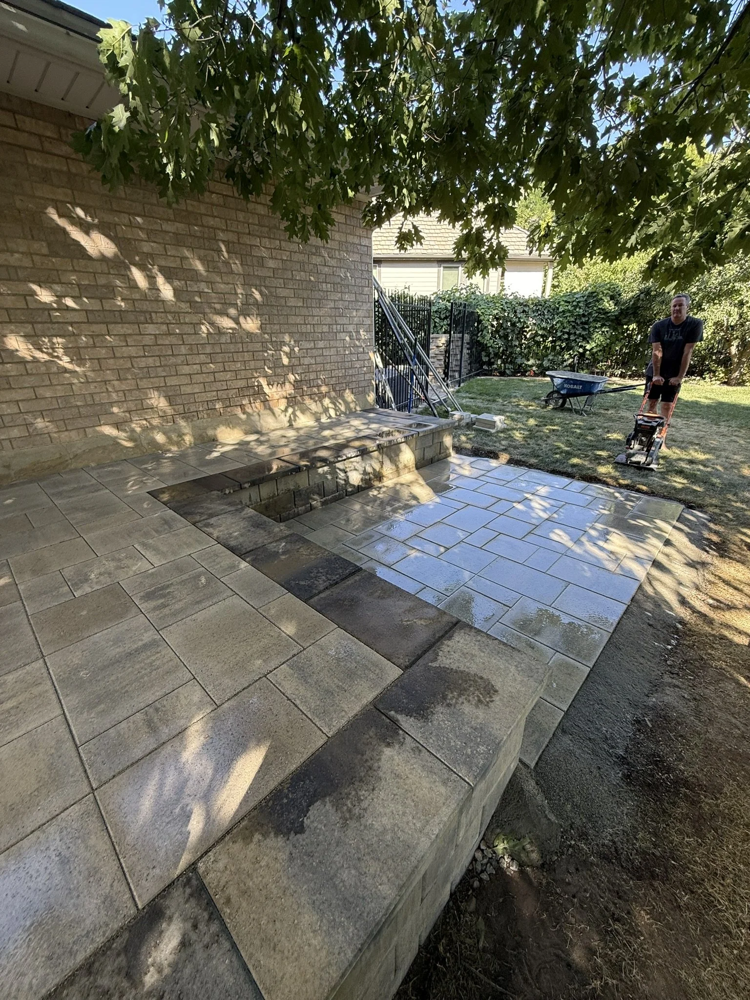 A man is working on a backyard patio made of stone tiles, with some tiles being wet, under a large leafy tree. There is a brick wall in the background, and a wheelbarrow and a power tool are also visible.