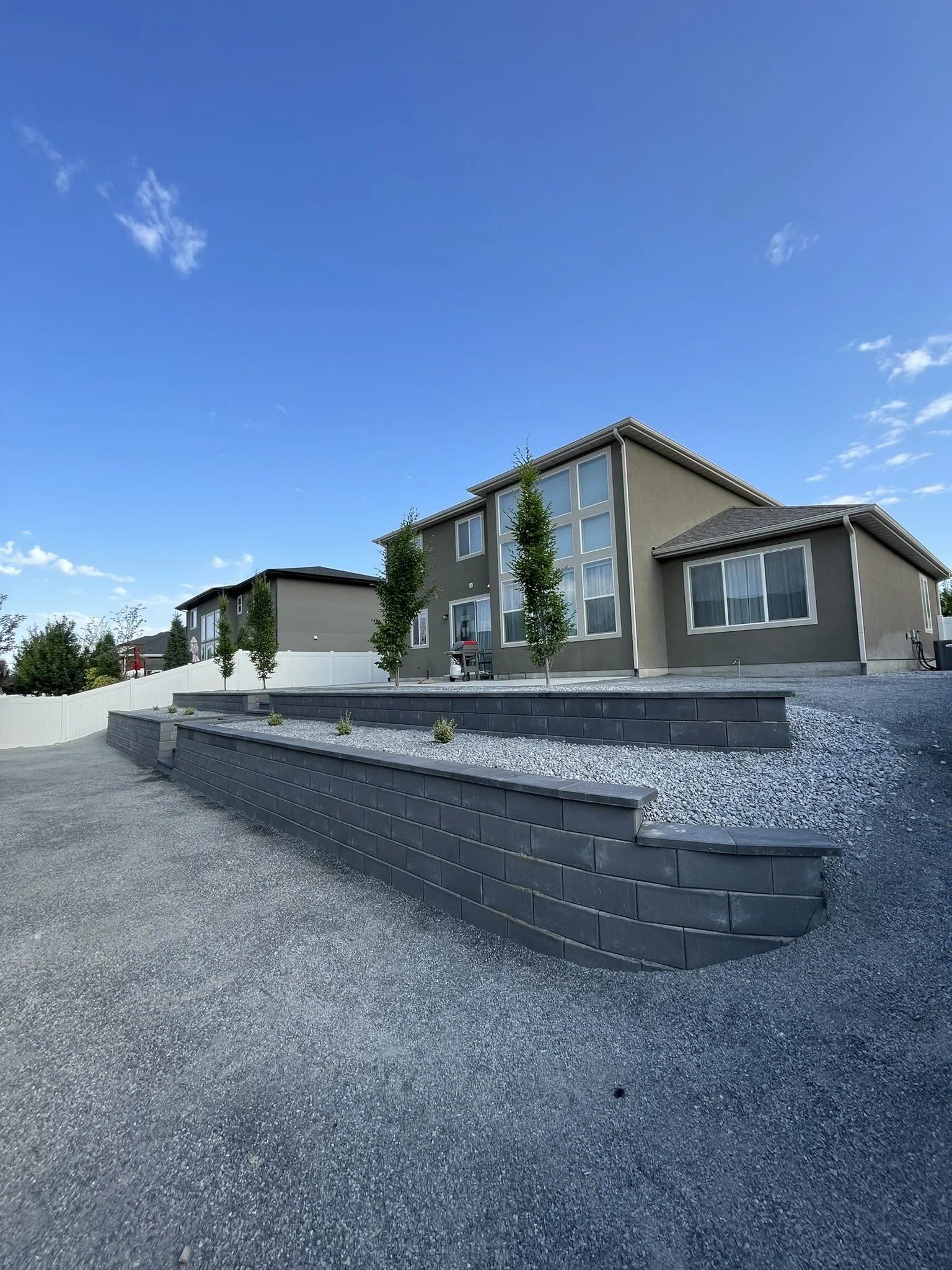 Modern two-story house with large windows, trees, and a tiered landscaped yard with gravel and retaining walls, under a clear blue sky.