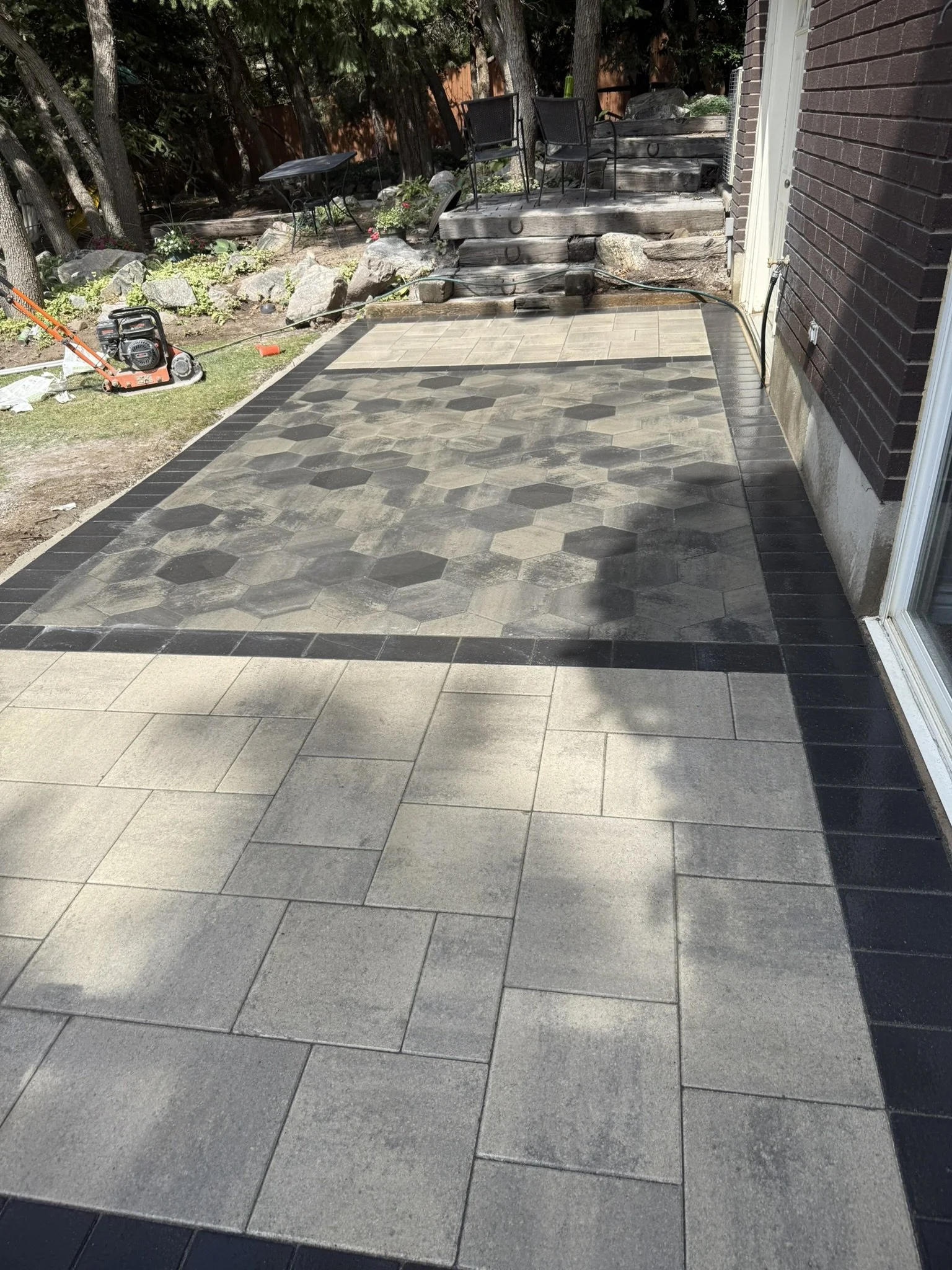 Backyard patio with freshly laid concrete tiles in a geometric pattern, bordered by black tiles, with garden chairs and a table on a wooden deck area with rocks and trees in the background.