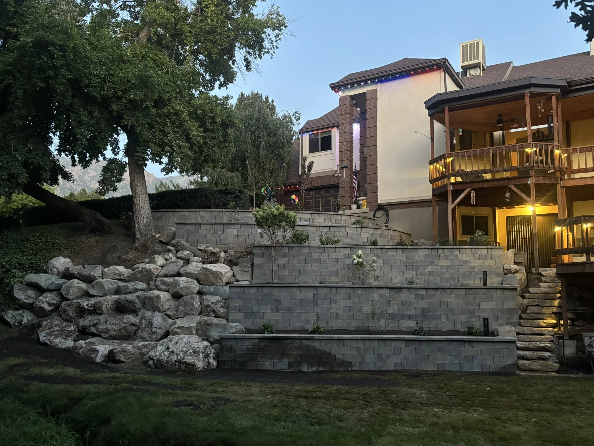 A backyard with tiered retaining walls made of grey concrete blocks, large rocks on one side, stairs leading to a house with a wooden deck on the upper level, surrounded by trees, with mountains visible in the background. The house has string lights 