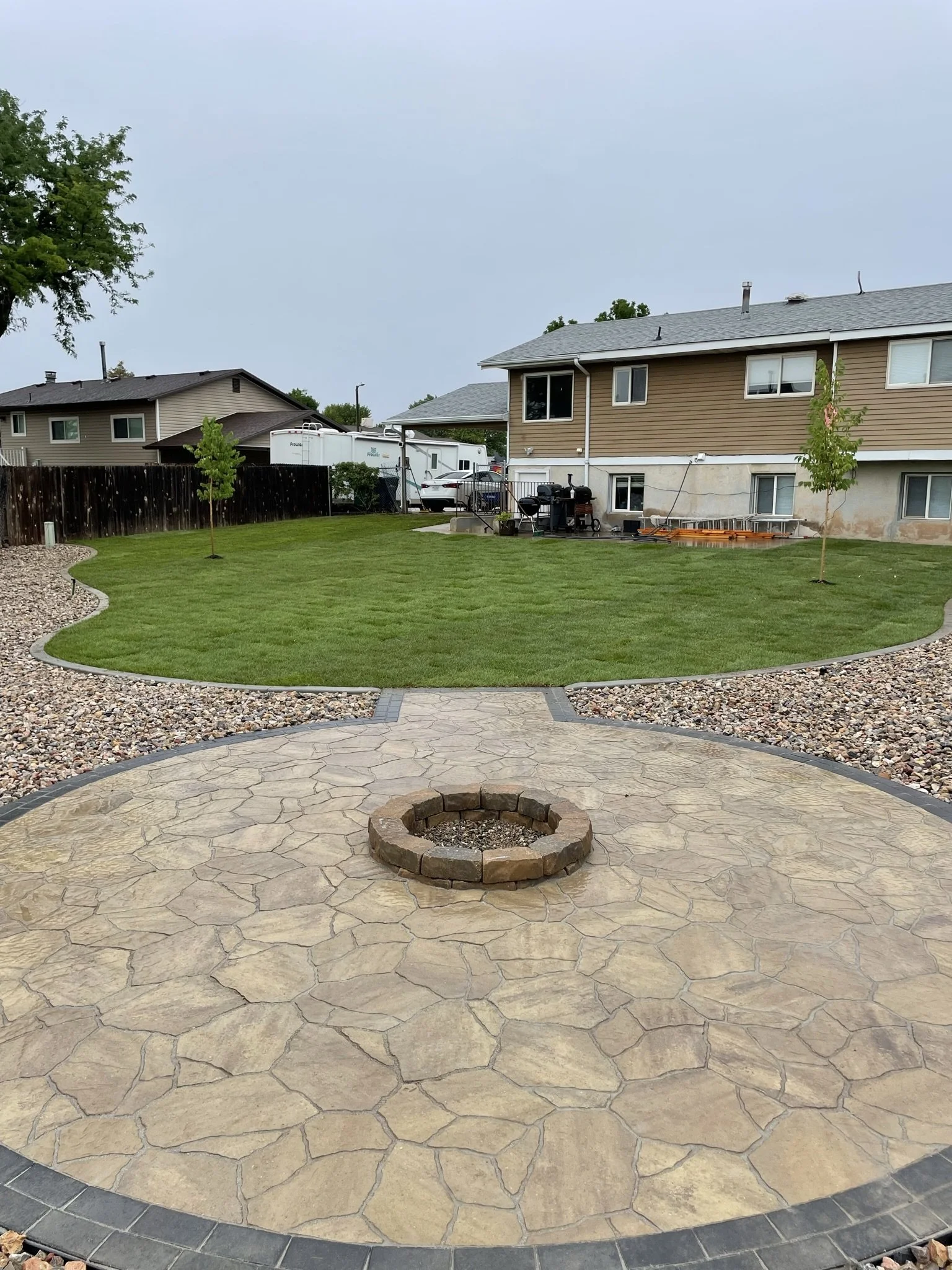 Backyard patio with a fire pit, stone pathway, green lawn, young trees, and neighboring houses with RVs and outdoor furniture.