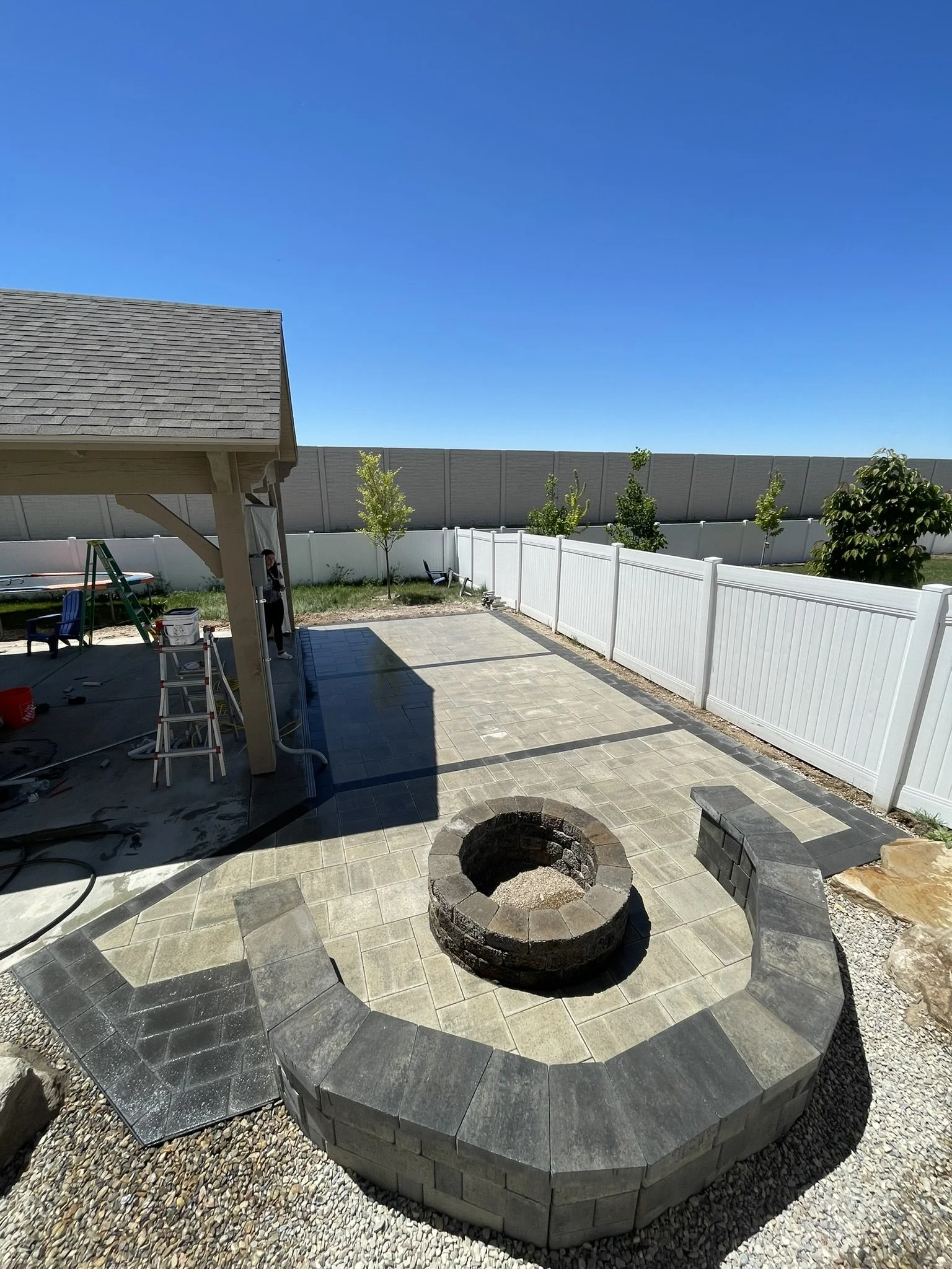 Backyard patio area with fire pit, white privacy fence, small trees, and construction work underway.