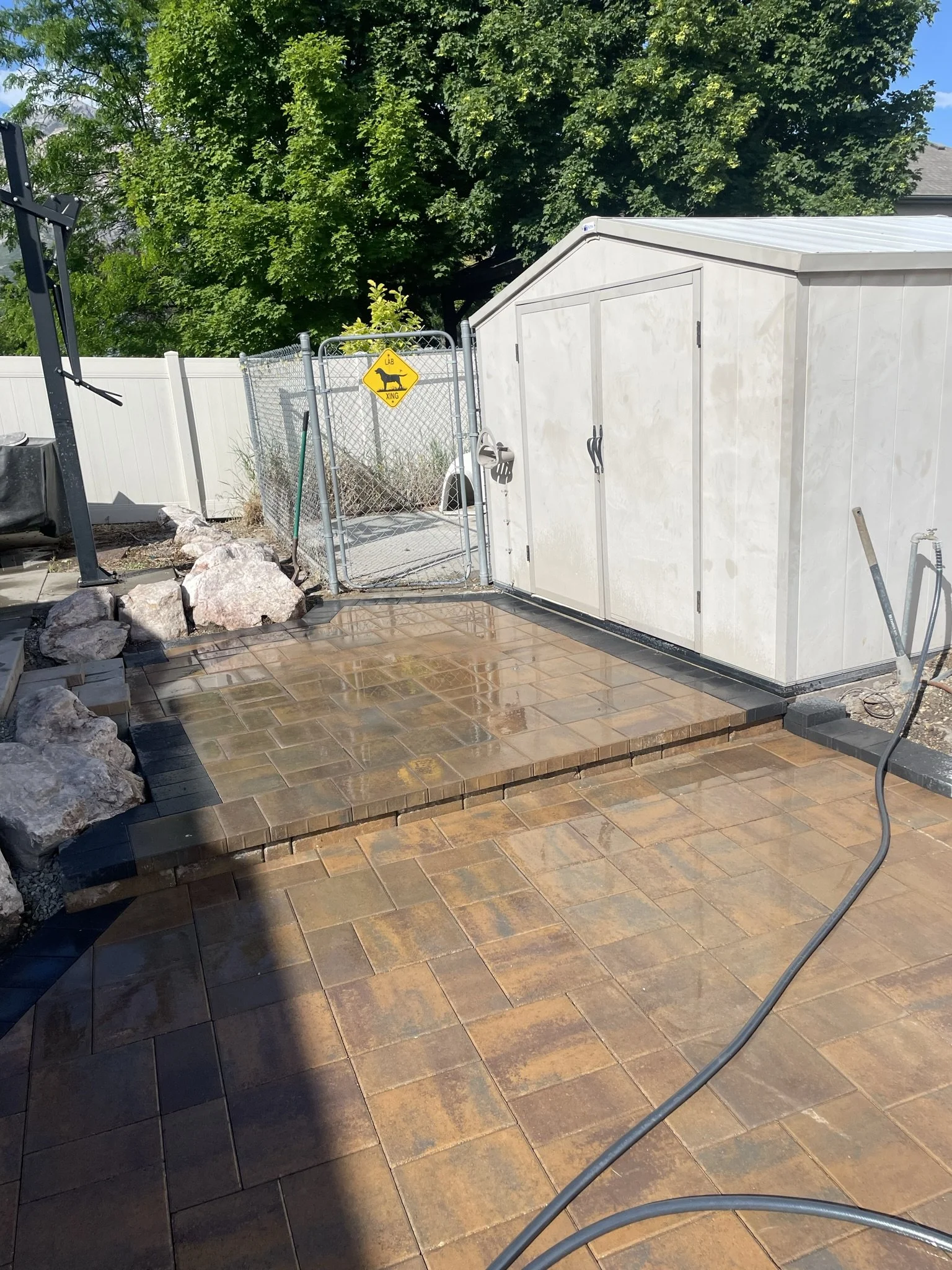 A backyard patio with wet, brown paving stones, a small raised platform, a chain-link fence gate with a yellow dog warning sign, a white storage shed, large rocks, a black outdoor furniture piece on the left, and a large green tree in the background.
