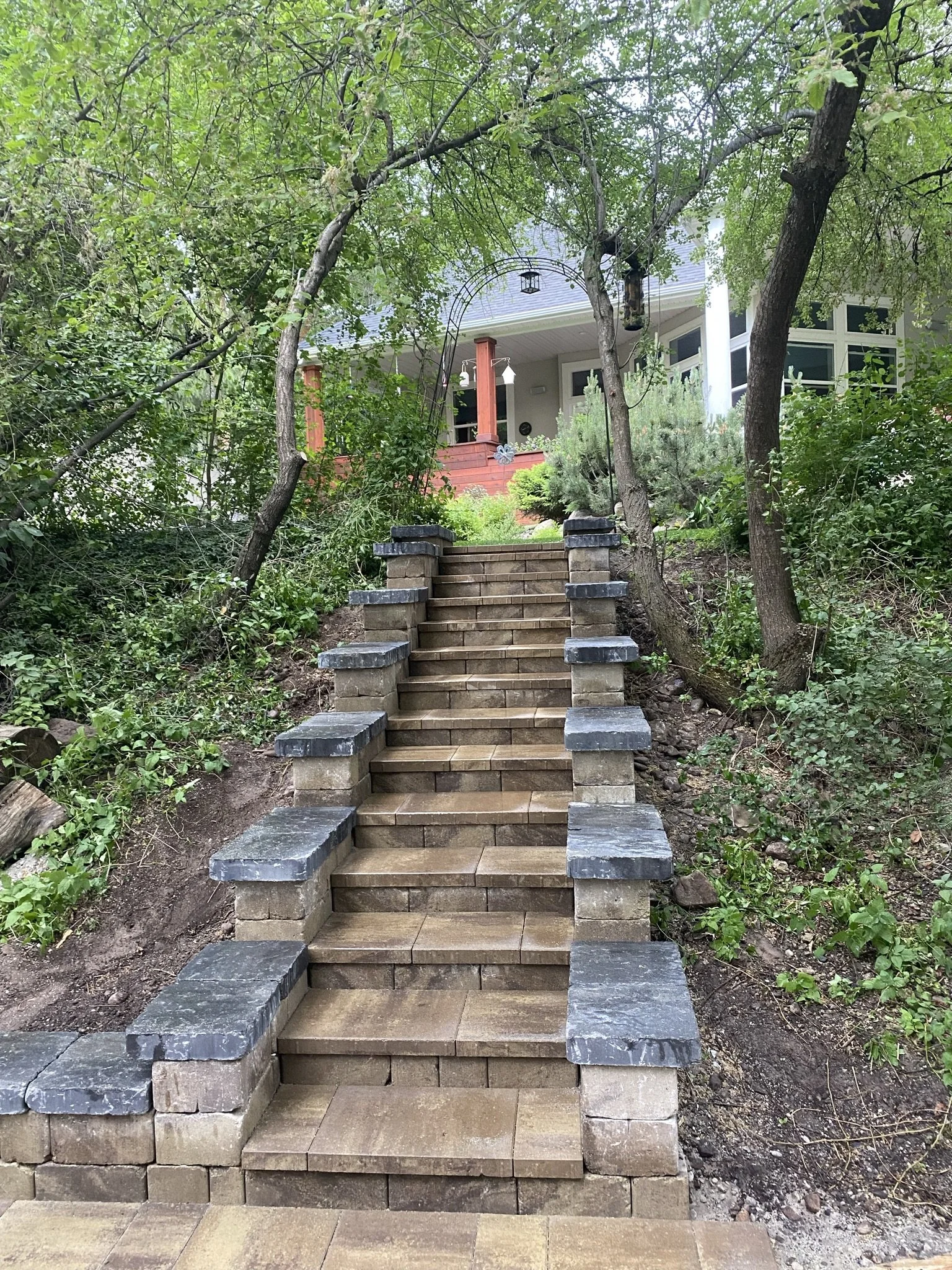 Stone staircase leading uphill to a house surrounded by greenery and trees, with a porch and hanging lantern.
