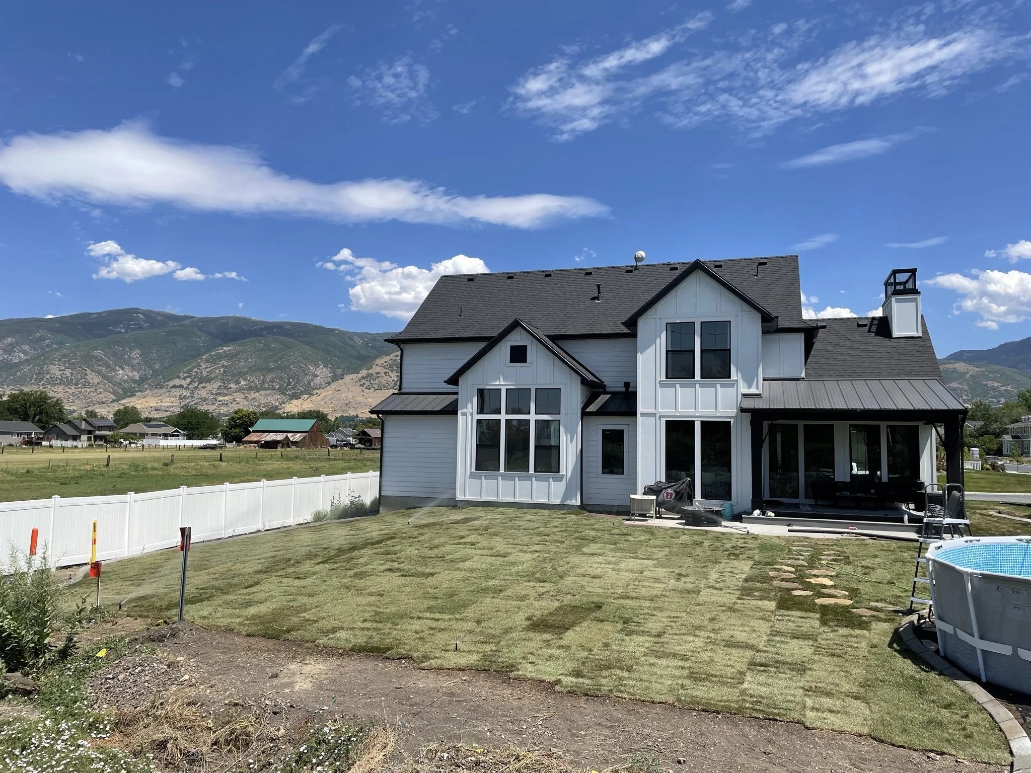 A two-story white house with black roof shingles, large windows, and a spacious backyard with a green lawn, a white fence, and an above-ground swimming pool, set against a backdrop of green mountains and blue sky with scattered clouds.