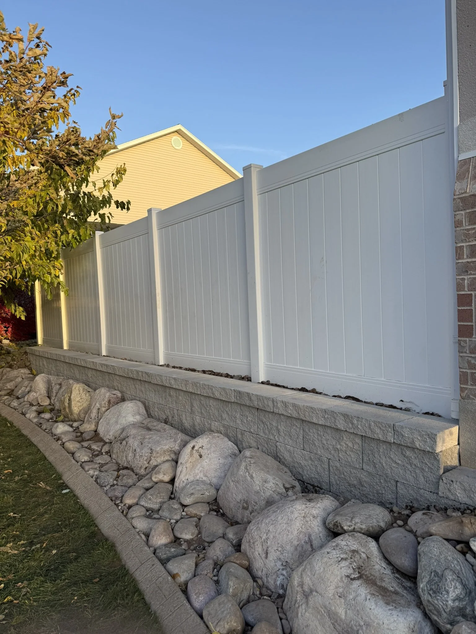 A white vinyl privacy fence on a stone retaining wall with large rocks and a landscaped border, next to a brick house with a guitar-shaped chimney pipe, under a blue sky.
