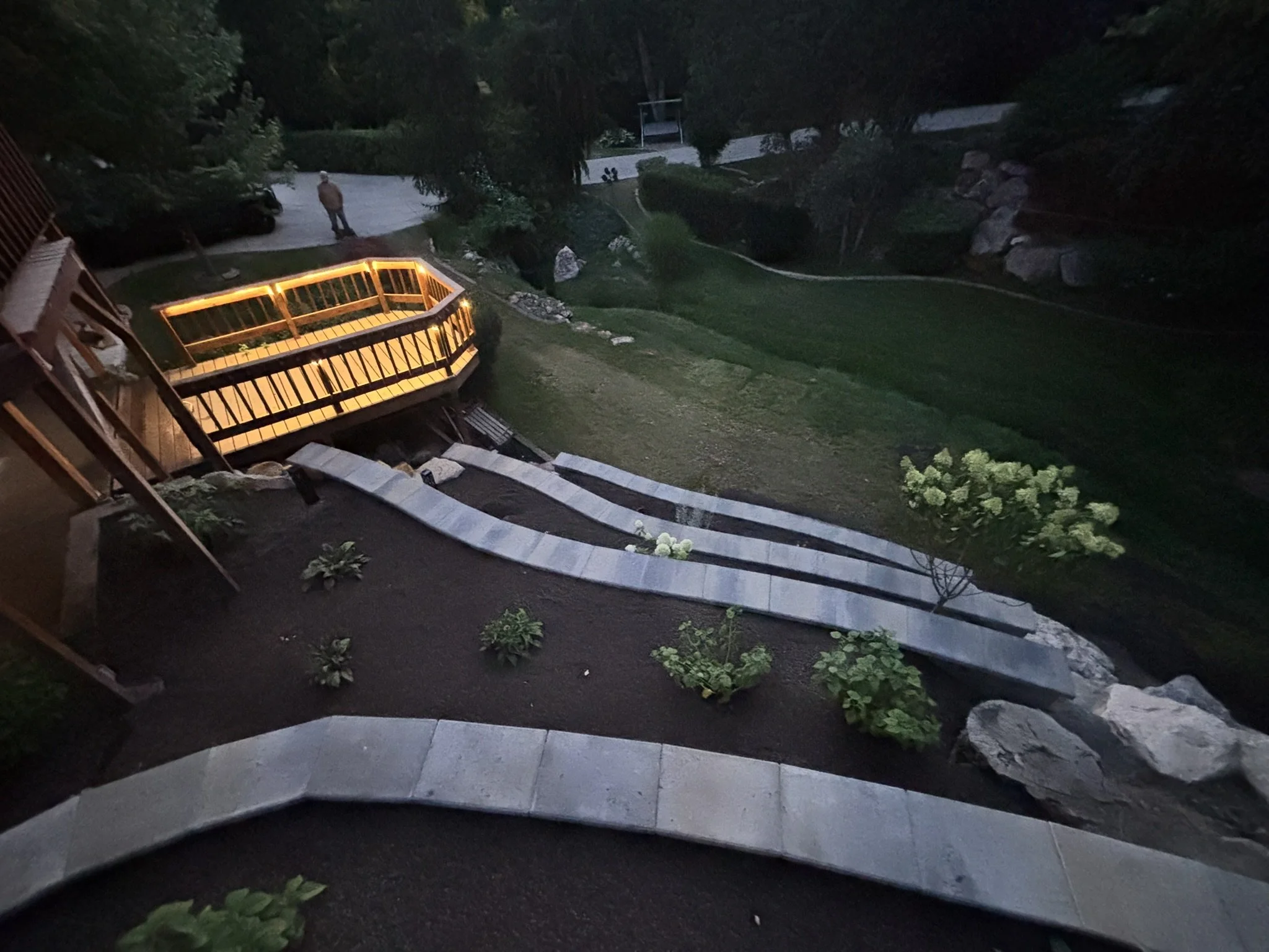 Nighttime view of a landscaped backyard with curved stone pathways, small shrubs, and a wooden deck with a lit railing, overlooking a grassy yard and trees.