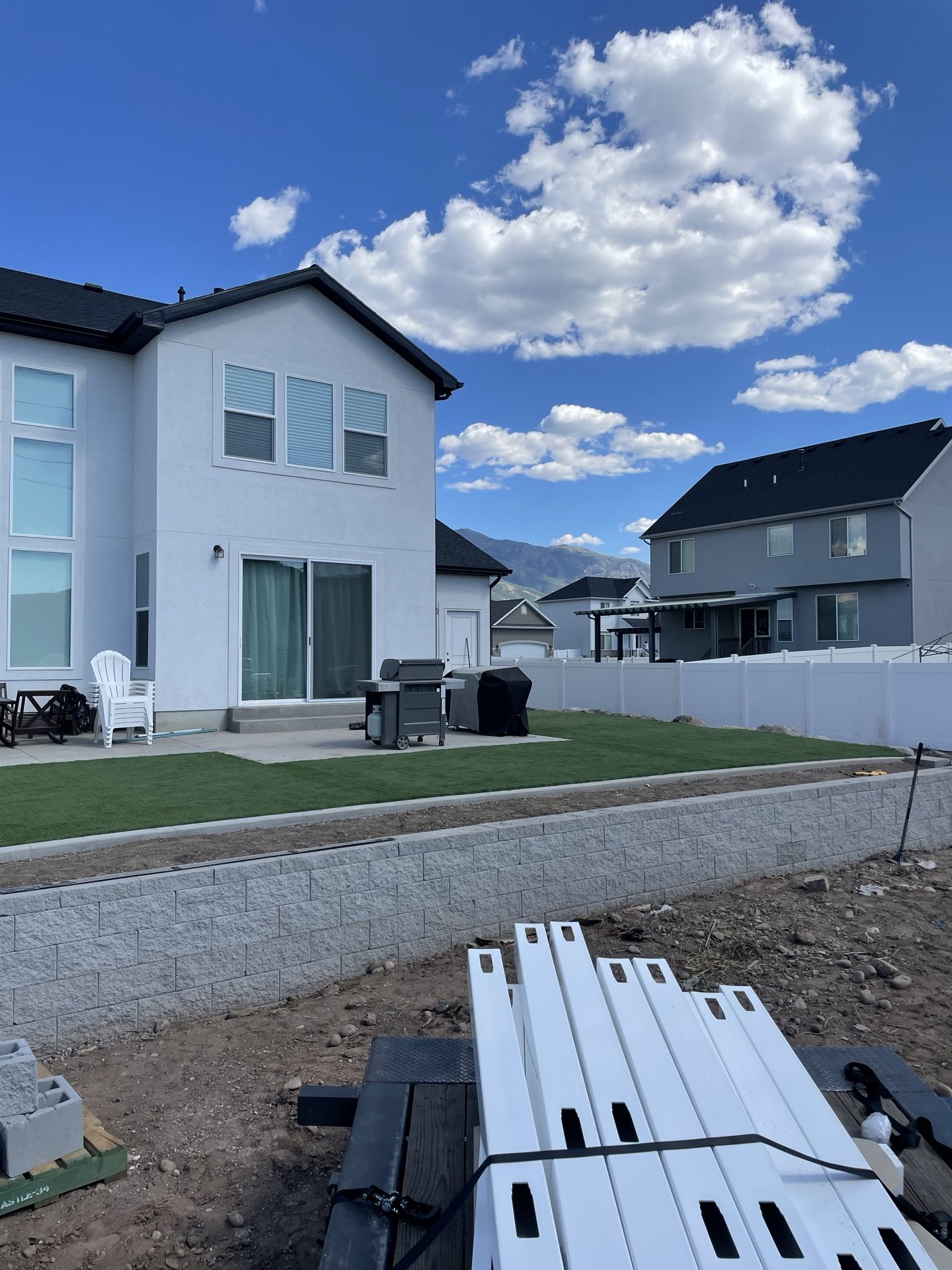 A backyard with a white house, a small patio, outdoor furniture, a barbecue grill, and a white fence, with mountains and a partly cloudy sky in the background.