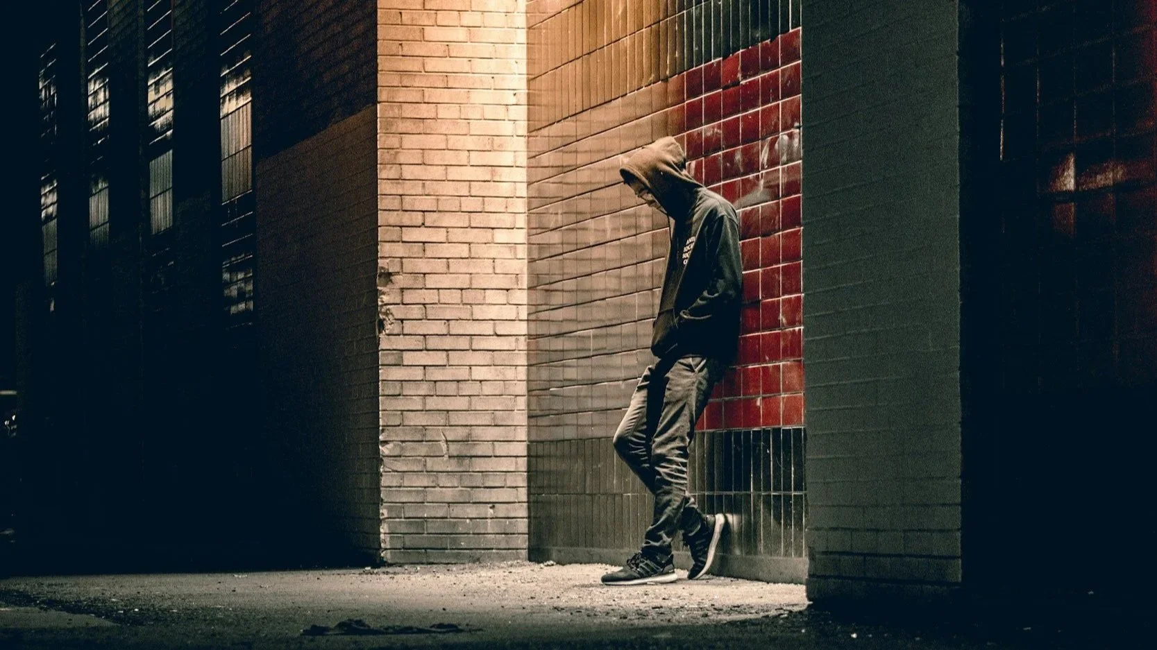 A person wearing a hoodie and jeans leaning against a brick wall at night, with shadows from nearby windows cast on the wall.