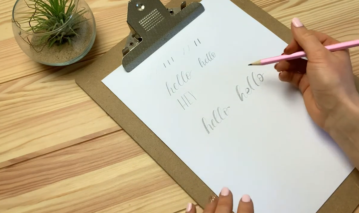 A close-up of a hand drawing in thick lines alongside the downstrokes of the word "hello" to create a faux calligraphy effect