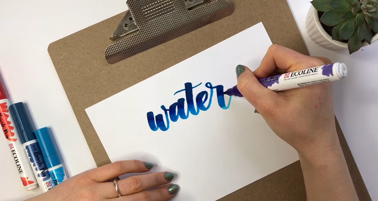 Purple being blended in: "Ultramarine violet Ecoline ink being blended into blue hand-lettered calligraphy, showing the three colours beginning to merge on textured paper."