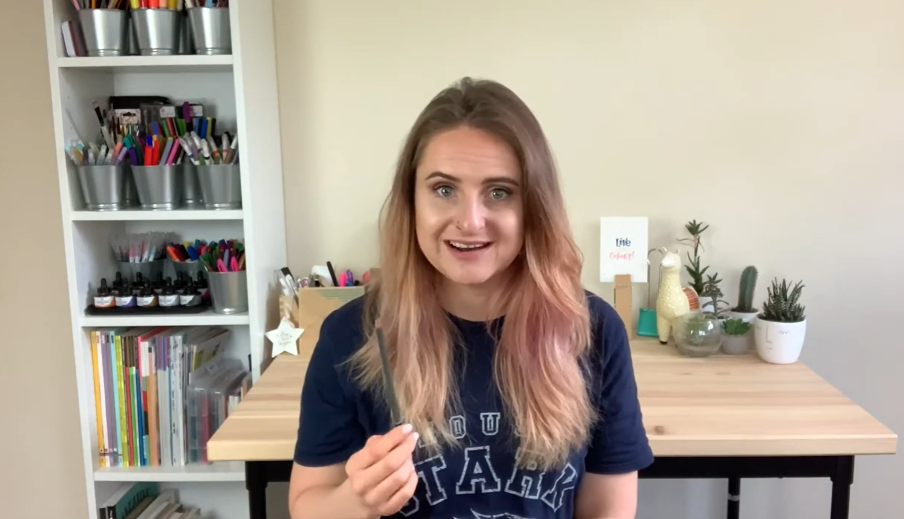 Milly, calligraphy tutor and founder of Blink Lettering, sitting at her desk holding a pencil and smiling at the camera