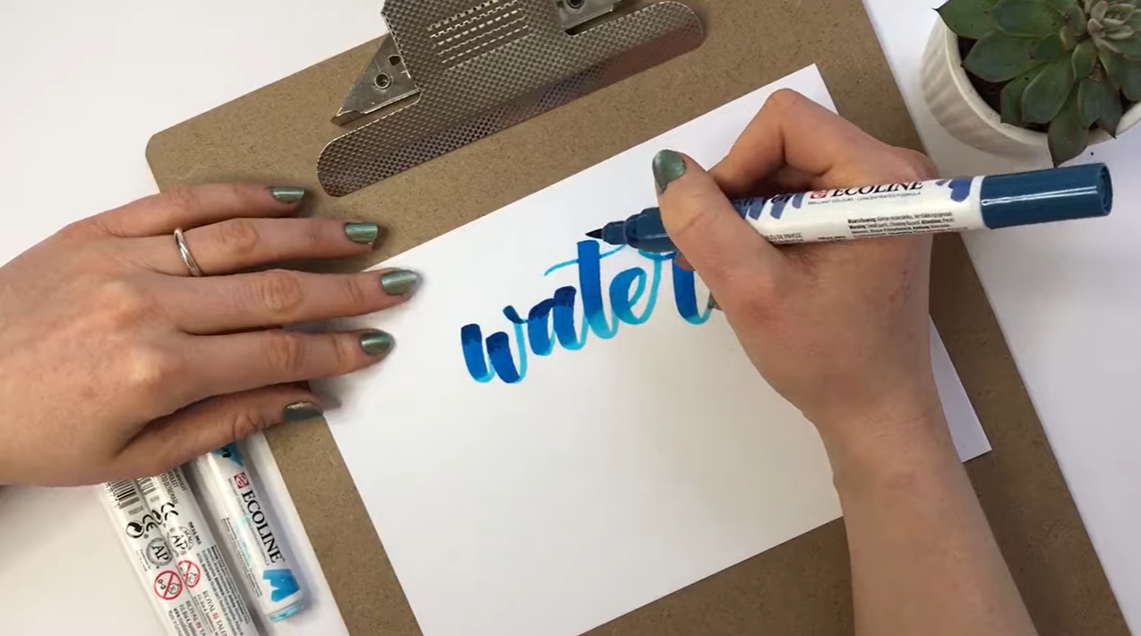 Light-to-dark blending in progress: "A cerulean blue Ecoline brush pen being lightly applied to the top of blue hand-lettered text, beginning the process of blending from light to dark.”