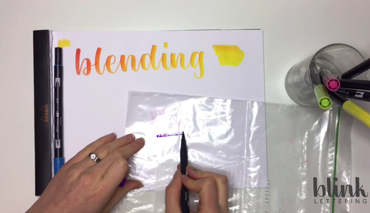 A sandwich bag with pink and purple Tombow ink coloured onto its surface, with brush pens laid alongside