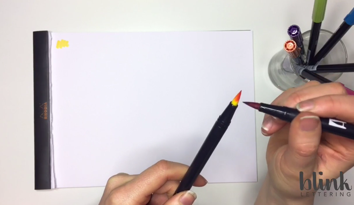 A close-up of a pink Tombow brush pen colouring directly onto the nib of a yellow Tombow pen to create a blended ink effect