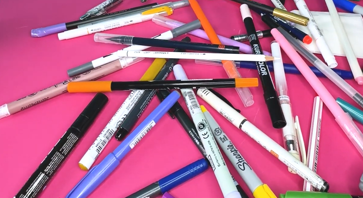A selection of calligraphy pens including flat-tipped nibs and brush pens laid out on a pink surface