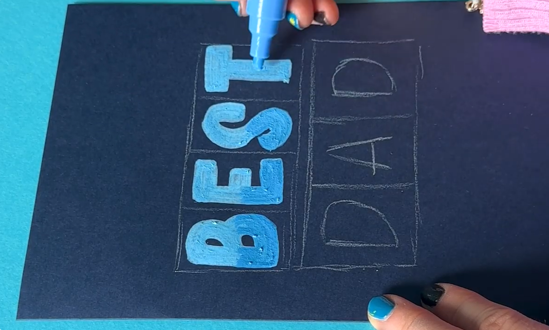 Bold uppercase letters being coloured with light blue and sky blue Posca paint markers, showing gradient shading applied towards the bottom of each letter for depth.