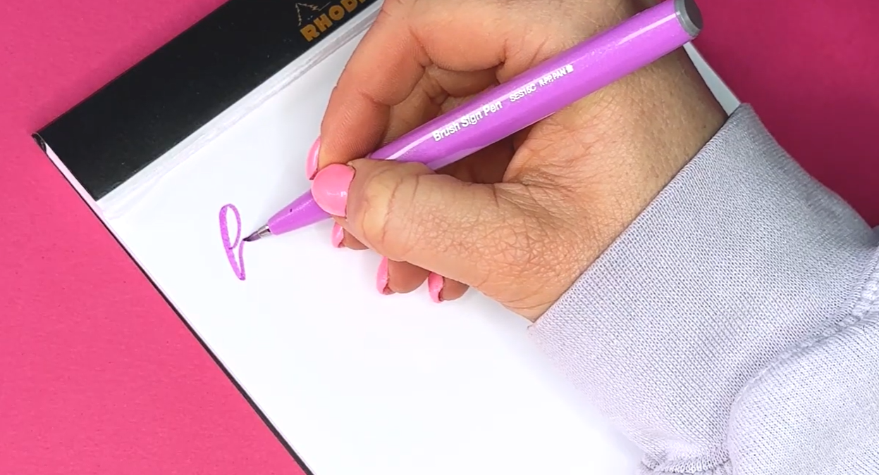 A hand holding a Pentel Brush Sign Pen, demonstrating the small flexible nib used for modern calligraphy