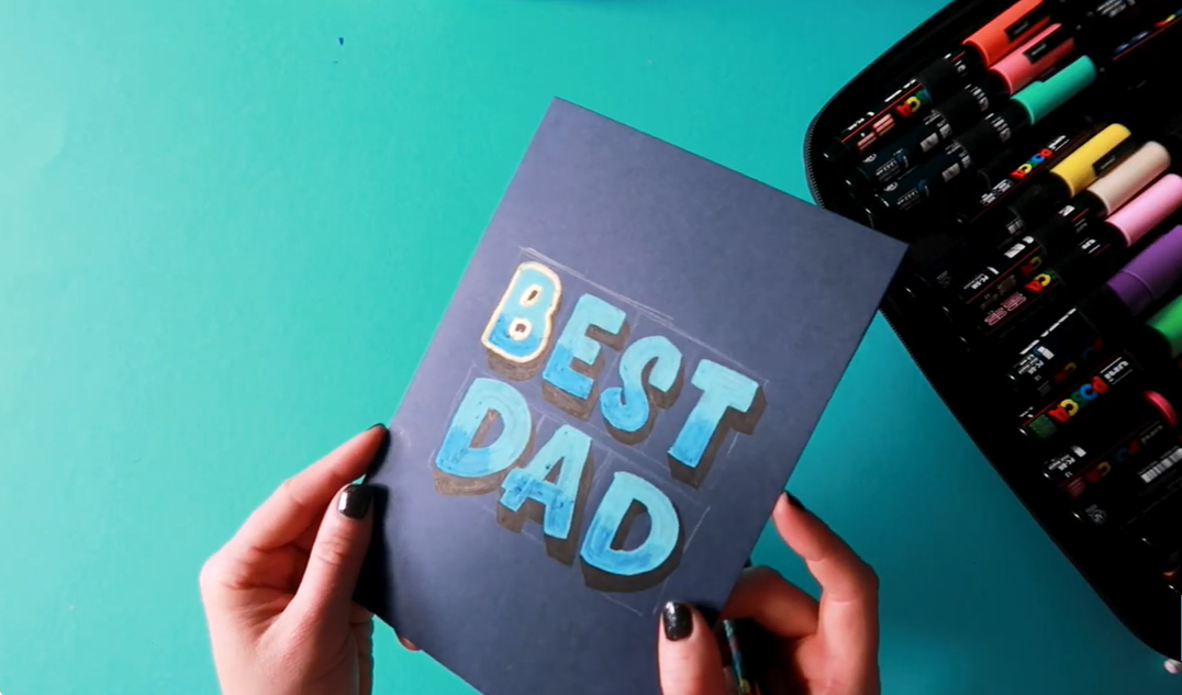 A gold Posca paint marker being used to outline bold blue hand-lettered text, creating a striking metallic border that makes the letters stand out against the dark card.