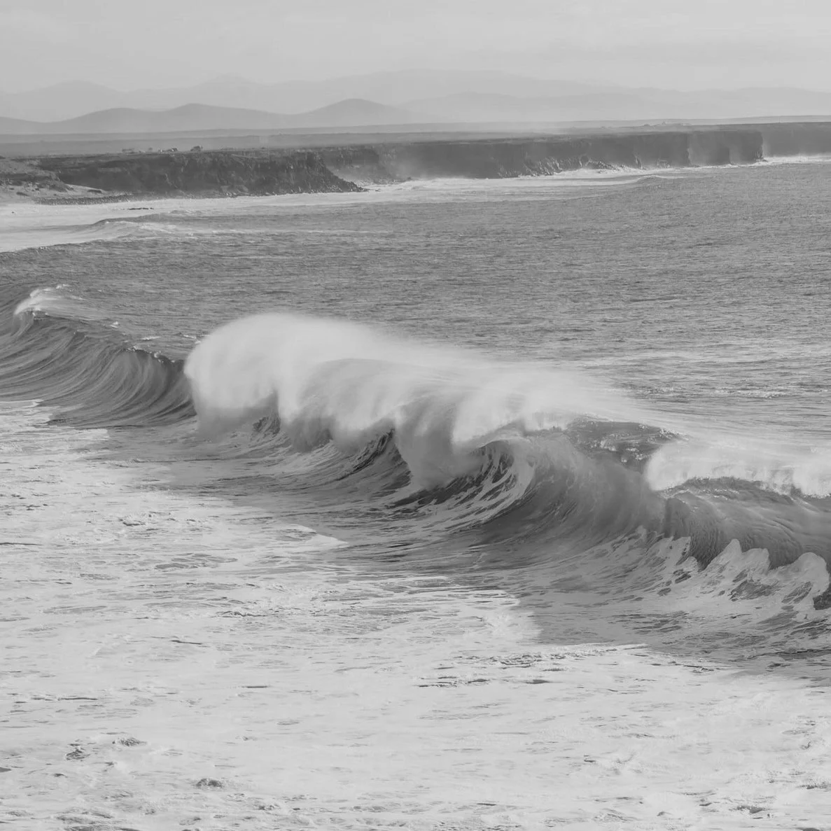 Black and white photograph of ocean waves crashing near a shoreline with cliffs in the background.