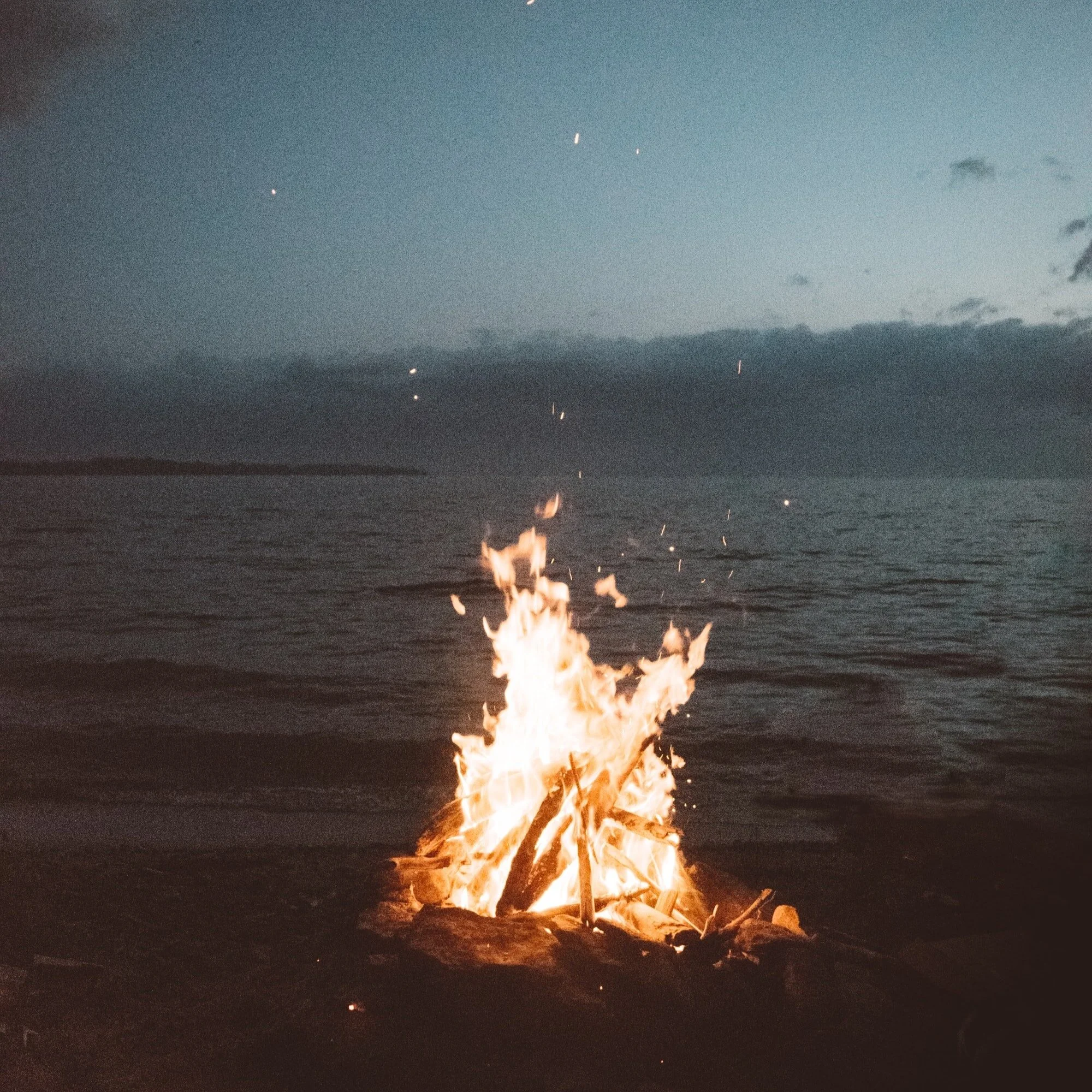 Bonfire burning on a beach during dusk with the ocean and cloudy sky in the background.