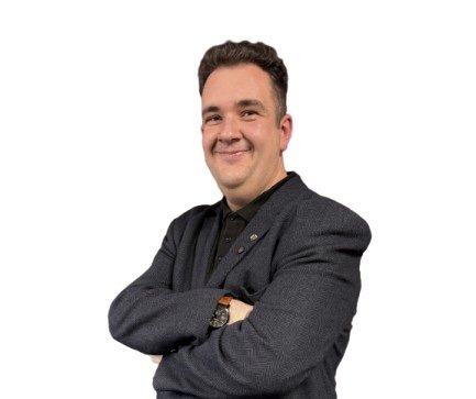 Professional headshot  an man smiling with arms crossed, wearing a charcoal grey blazer over a black polo shirt and a brown leather strap watch.