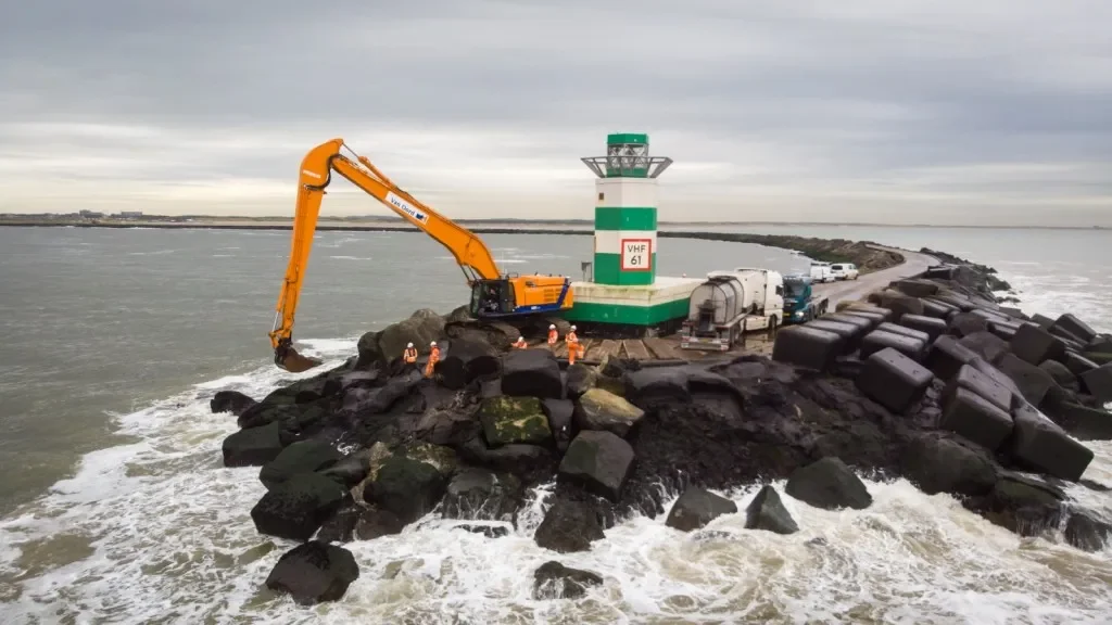 Een haven met een scheepswachttoren, een grote graafmachine die stenen verplaatst op een kunstmatige aanlegsteiger, en een groep mensen in oranje veiligheidskleding, tegen een skyline met bewolkte hemel.