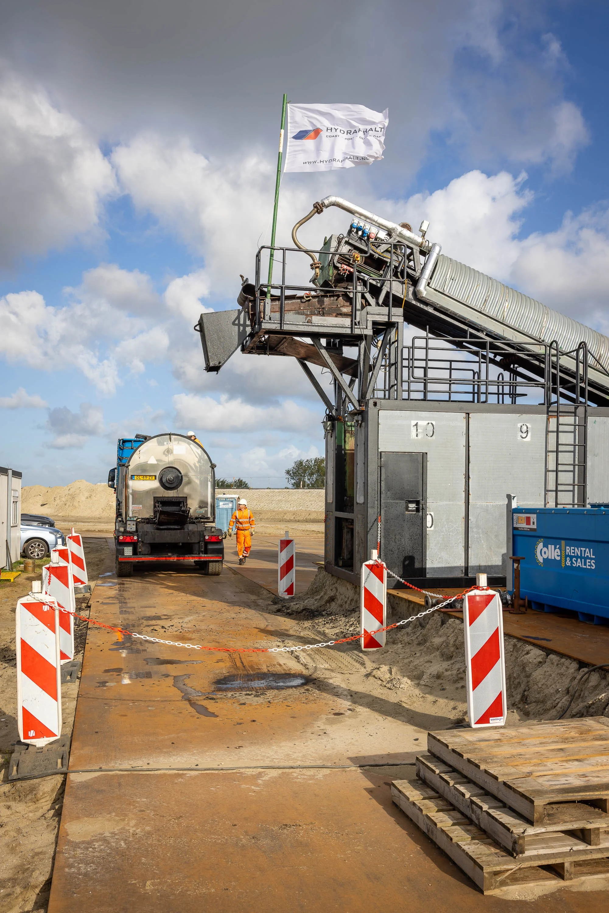 Werkzaamheden op een bouwplaats waarbij weg wordt aangelegd met beton. Er staan wegversperringen en er zijn werknemers in oranje veiligheidskleding.