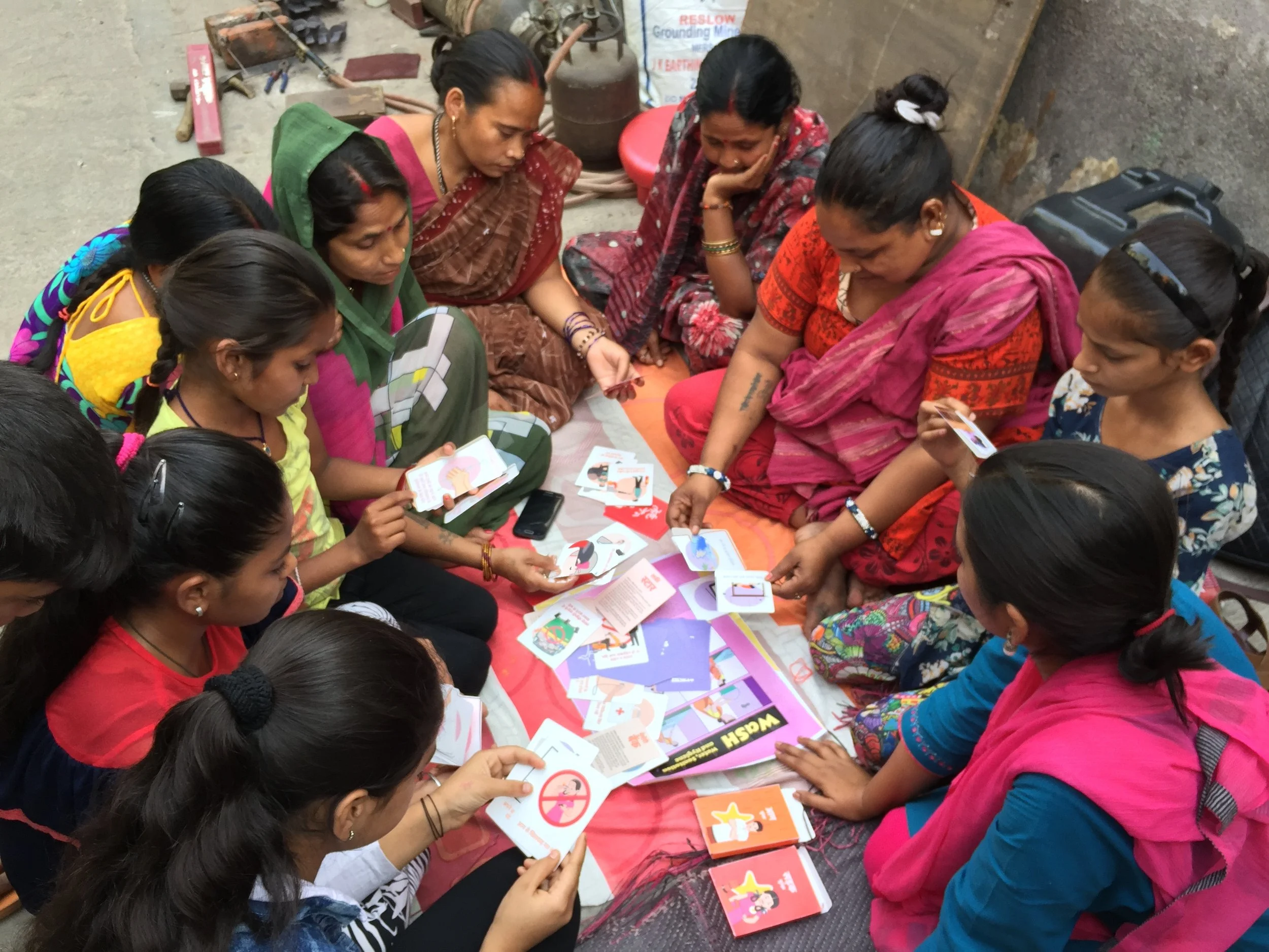 A group of women sitting on the ground, engaging in an educational session with colorful cards and a book spread on a mat. They appear to be discussing health or hygiene topics.