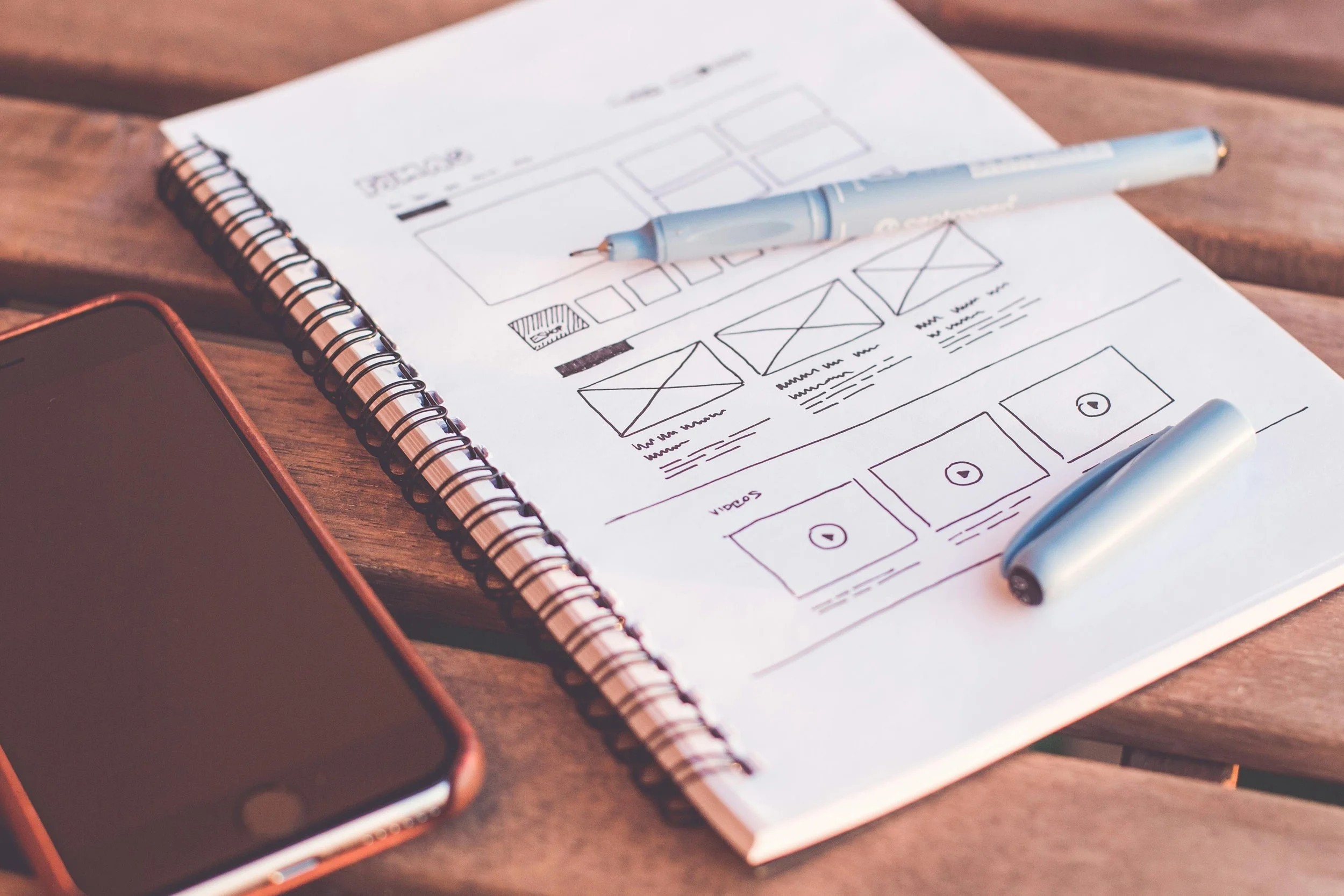 Notebook with website sketches, smartphone, blue pen on wooden table.