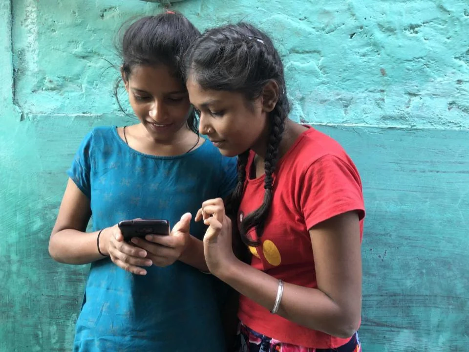 Two girls looking at a smartphone together against a turquoise wall.