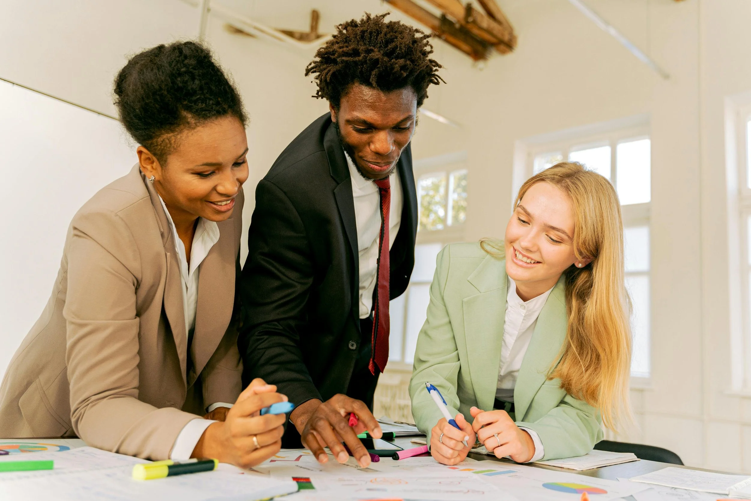 Three business professionals collaborating around a table with charts and graphs, using markers in an office setting.