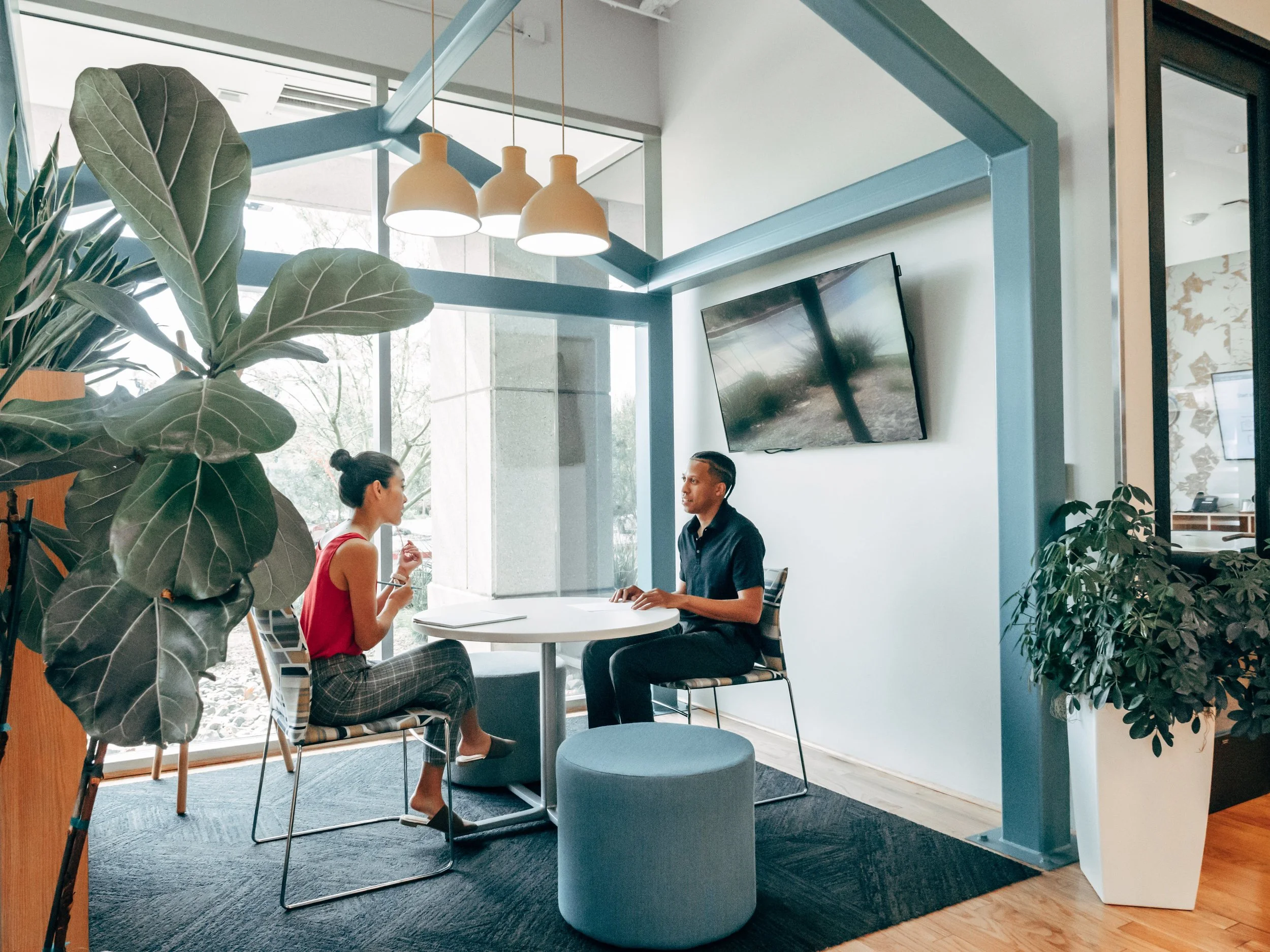 Two people sitting and talking at a table in a modern office area with large plants, pendant lights, and a wall-mounted TV.