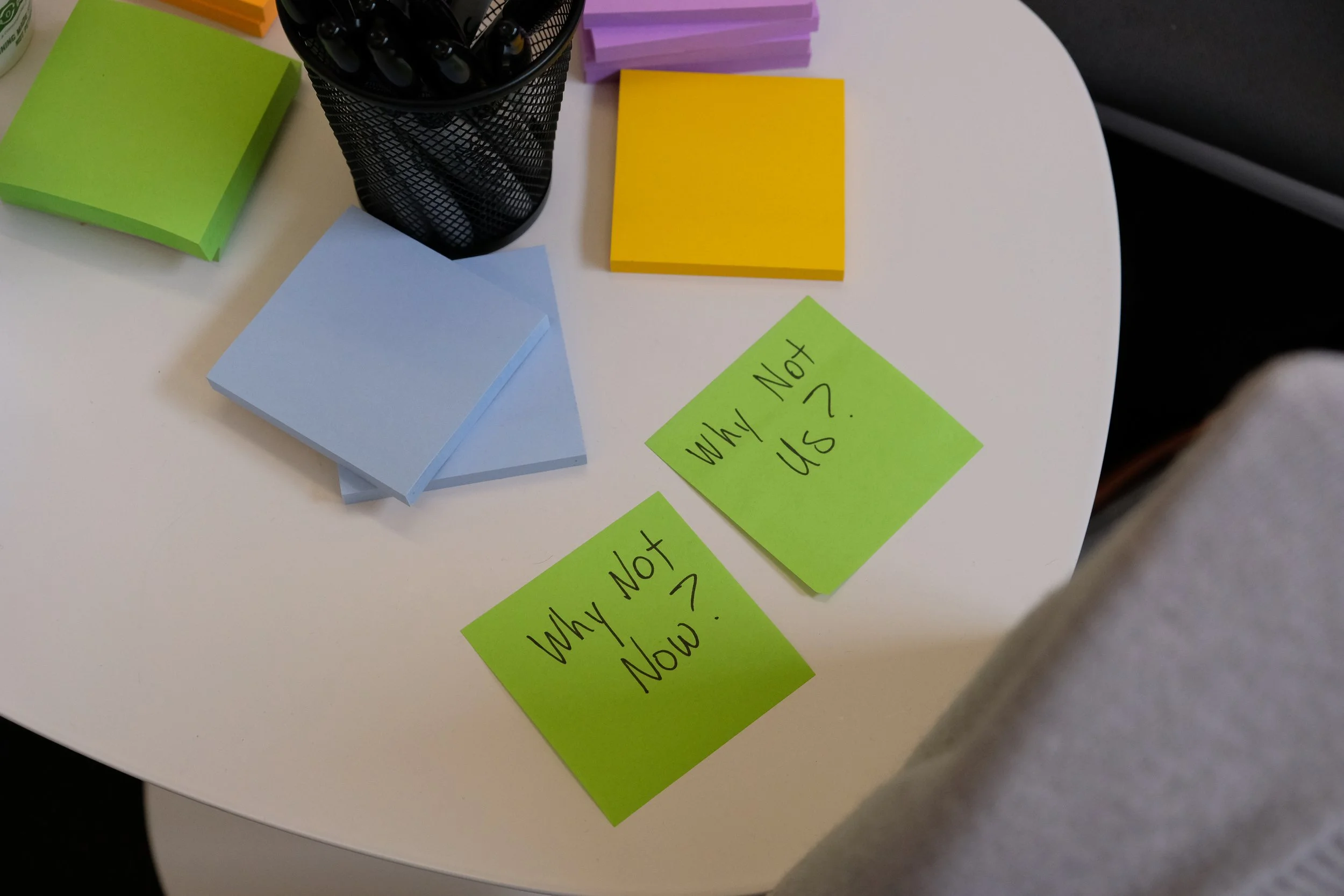Colorful sticky notes with questions 'Why Not Now?' and 'Why Not Us?' on a white table, with colored paper stacks and a black pen holder visible.