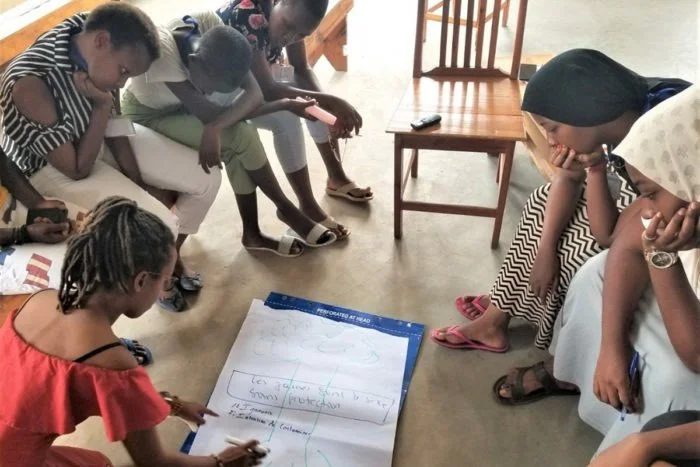 A group of people sitting around a large sheet of paper on the floor, engaging in discussion and note-taking. The scene appears to be a collaborative brainstorming or workshop session in a casual setting.