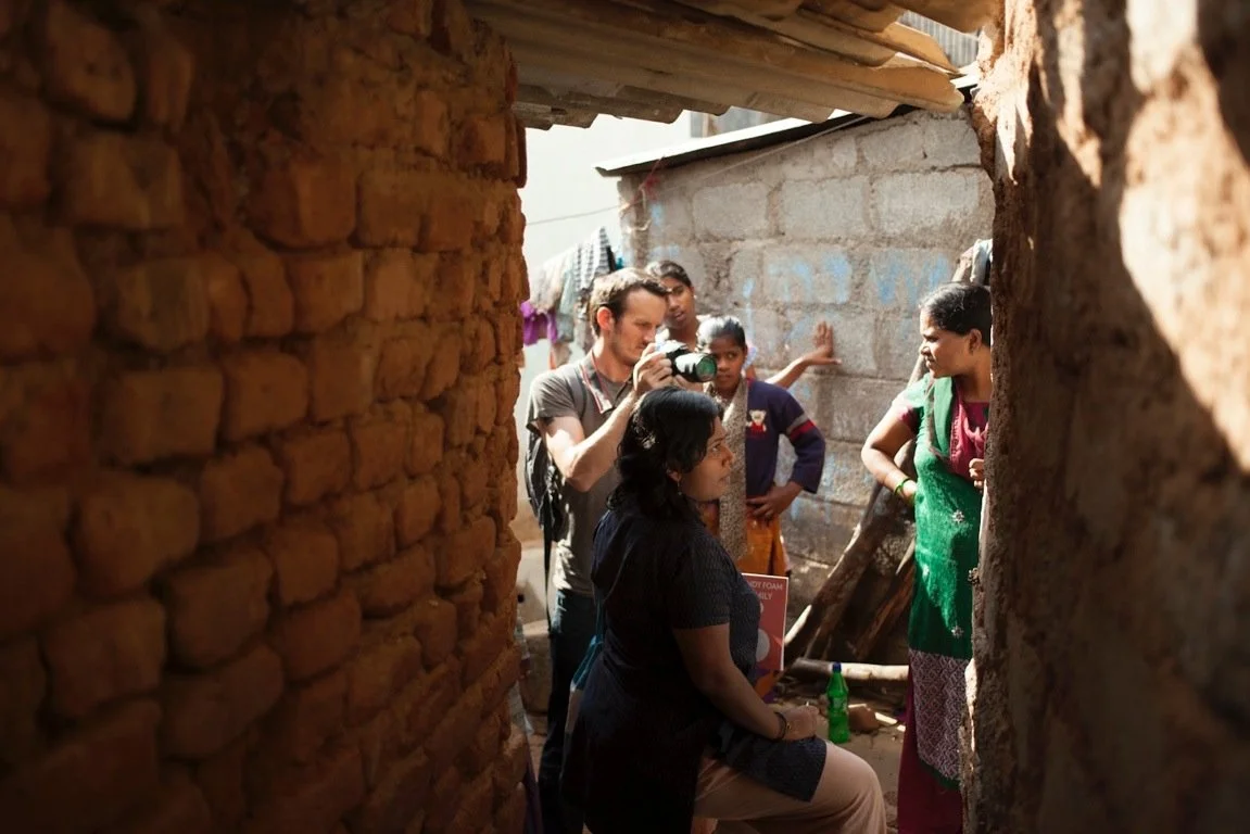 Group of people in a narrow alley with brick walls, one holding a camera, others engaging in conversation.