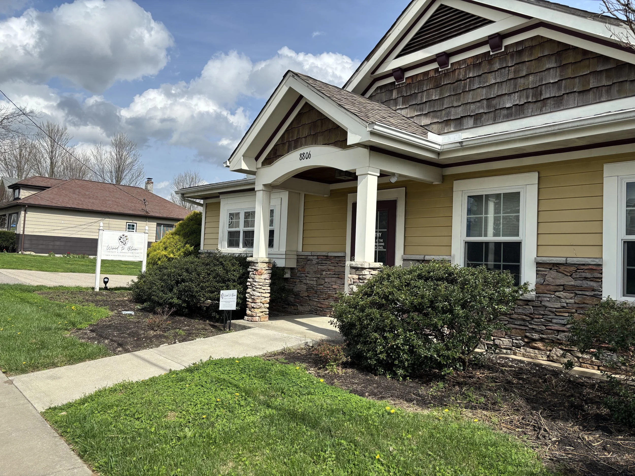 A yellow Craftsman-style house with stone accents on the porch and a gabled roof, surrounded by green bushes and a neatly maintained lawn, under partly cloudy skies.
