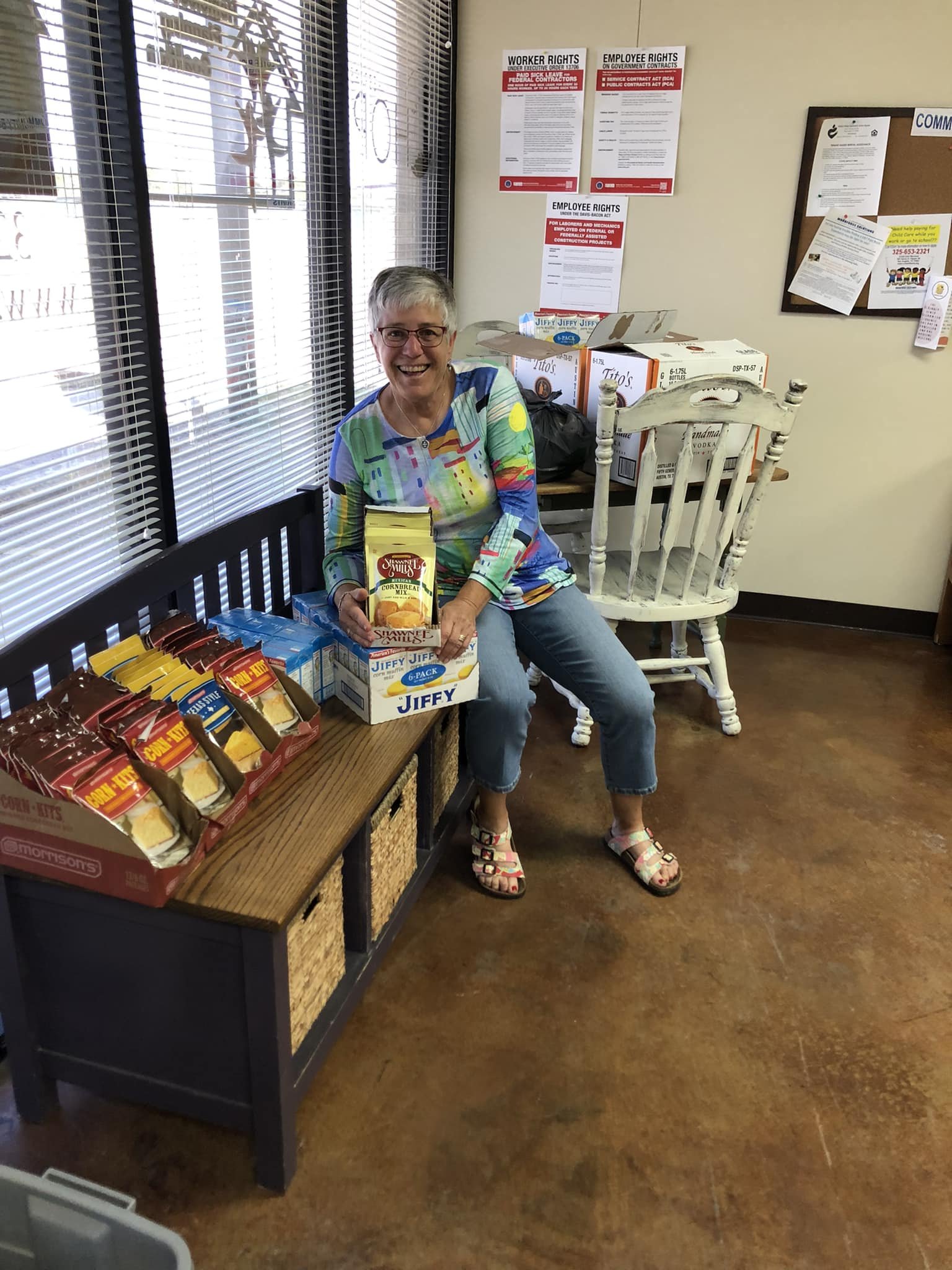 A smiling woman sitting on a bench holding a box of cornbread mix, surrounded by boxes of Jiffy cornbread mix and other food items in a community center or food bank. There are informational posters on the wall behind her and a window with blinds to 
