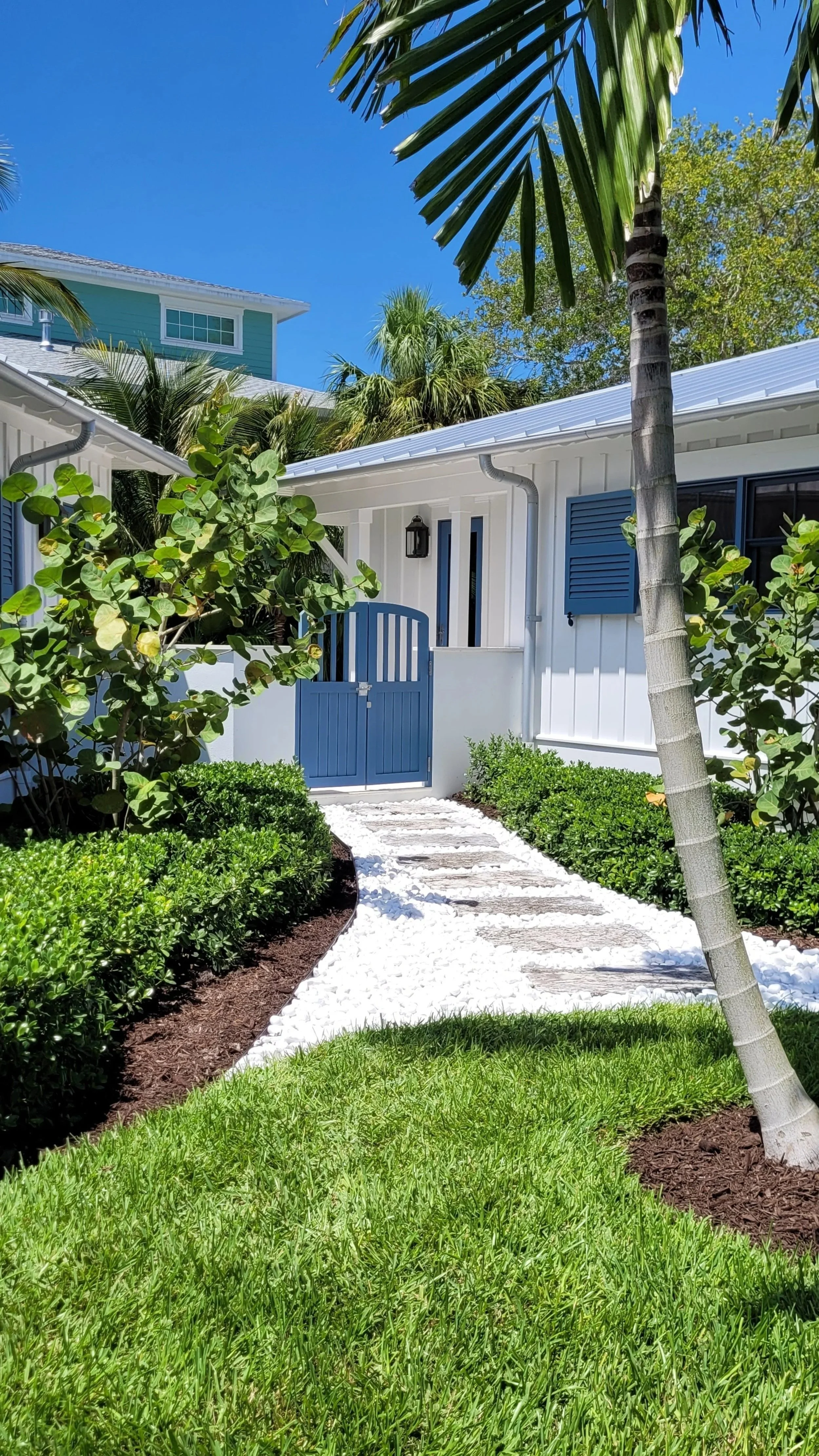 A white house with blue shutters and a blue gate, surrounded by tropical plants and trees, under a clear blue sky.