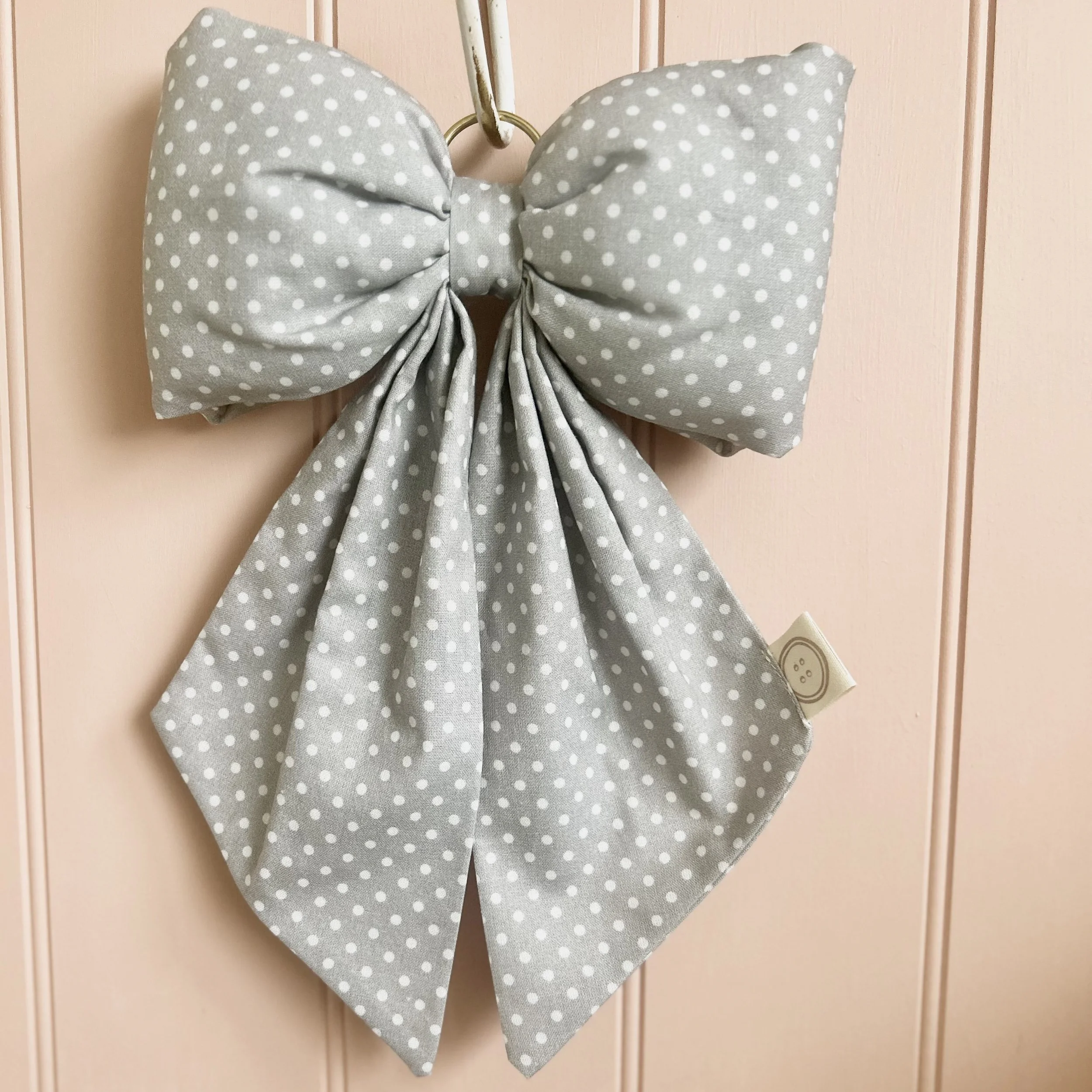 Blue/grey polka dot fabric bow hanging from a hook on a pale pink shiplap background.. Perfect for nursery decor