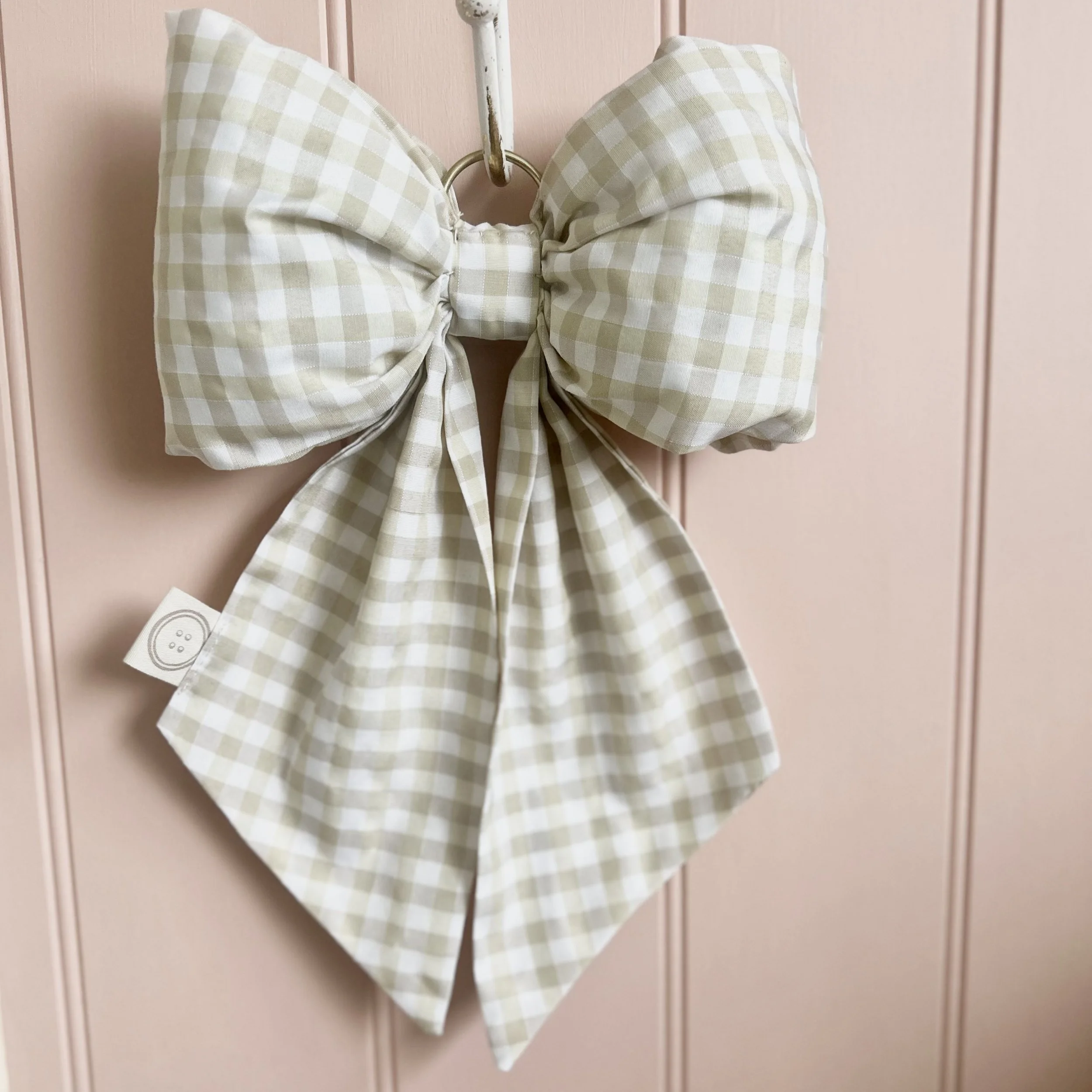 Cream gingham fabric bow hanging from a hook on a pale pink shiplap background.. Perfect for nursery decor