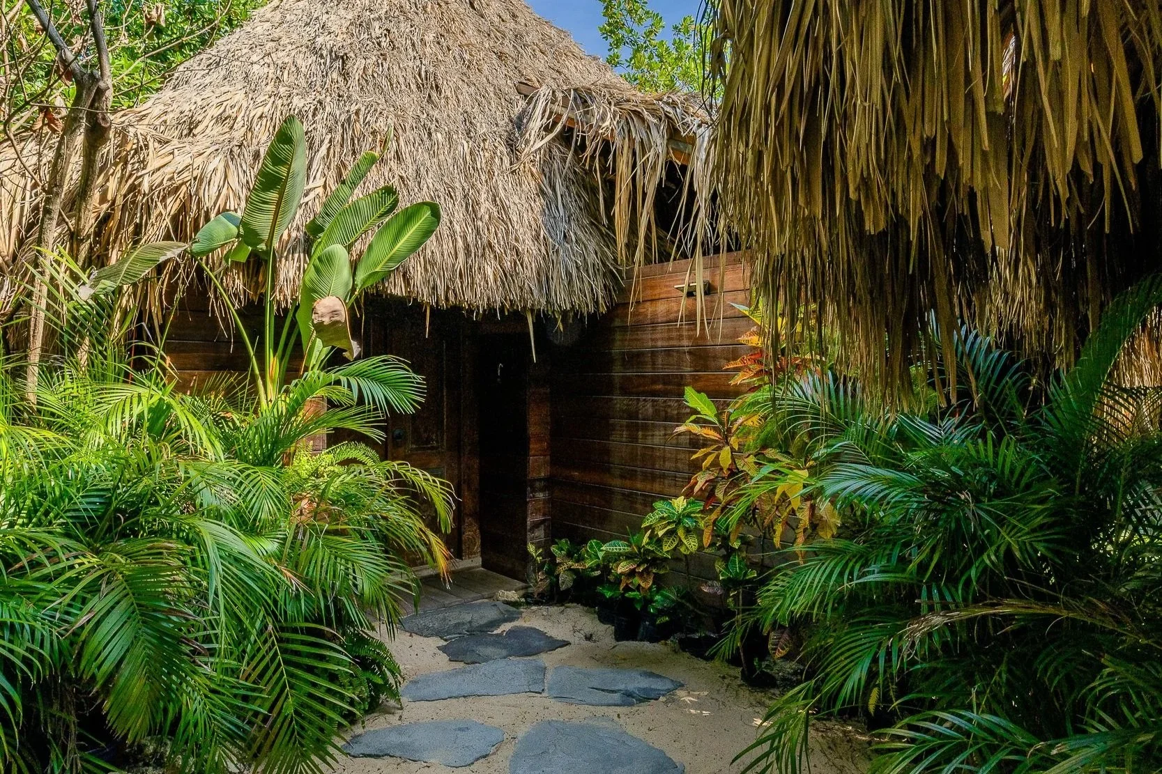 Beachside tiki hut with thatched roof, surrounded by tropical plants, palm trees, and sand, with a pink chair inside the hut.