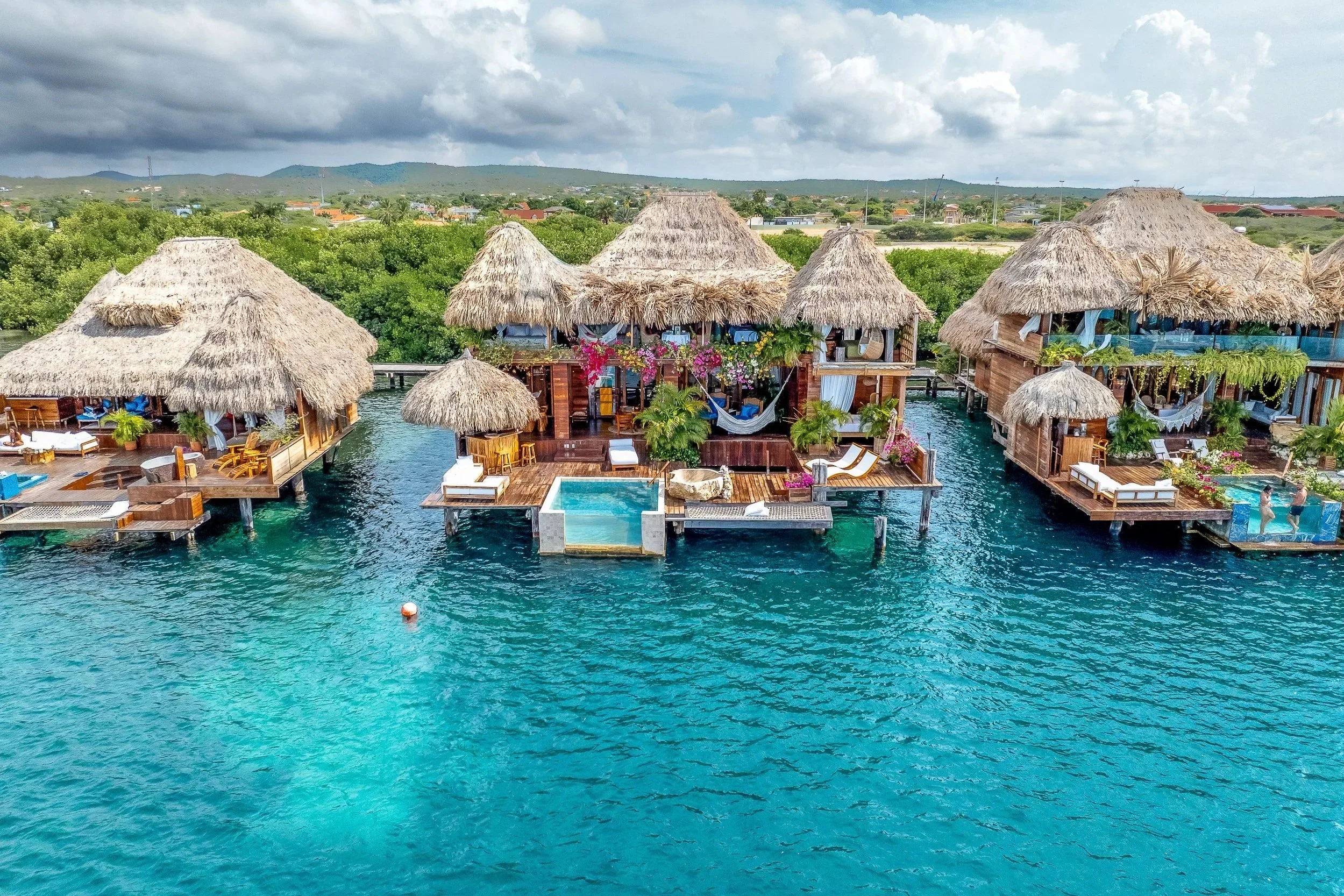 Overwater bungalow with thatched roofs on a tropical body of water, under a partly cloudy sky.