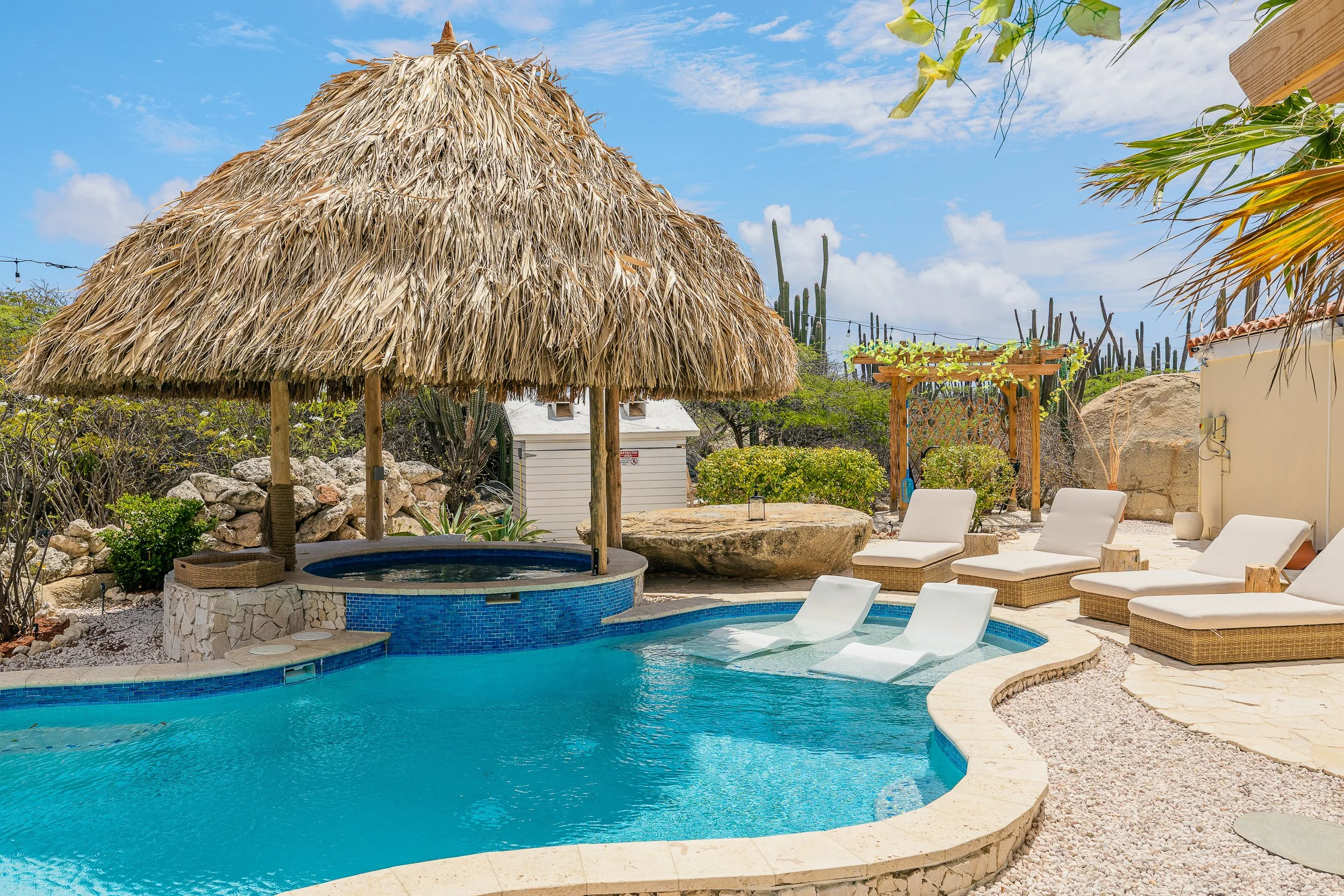 A house with a red tile roof, a backyard with a swimming pool, hot tub, two lounge chairs, and surrounding desert landscaping with cacti and green plants.