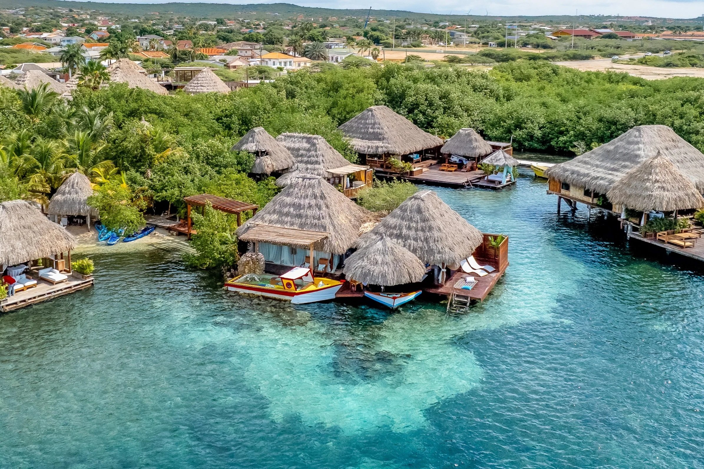 Overwater bungalows with thatched roofs, a swimming pool, lounge chairs, and palm trees along a sandy shore, with clear turquoise water and a boat in the distance.