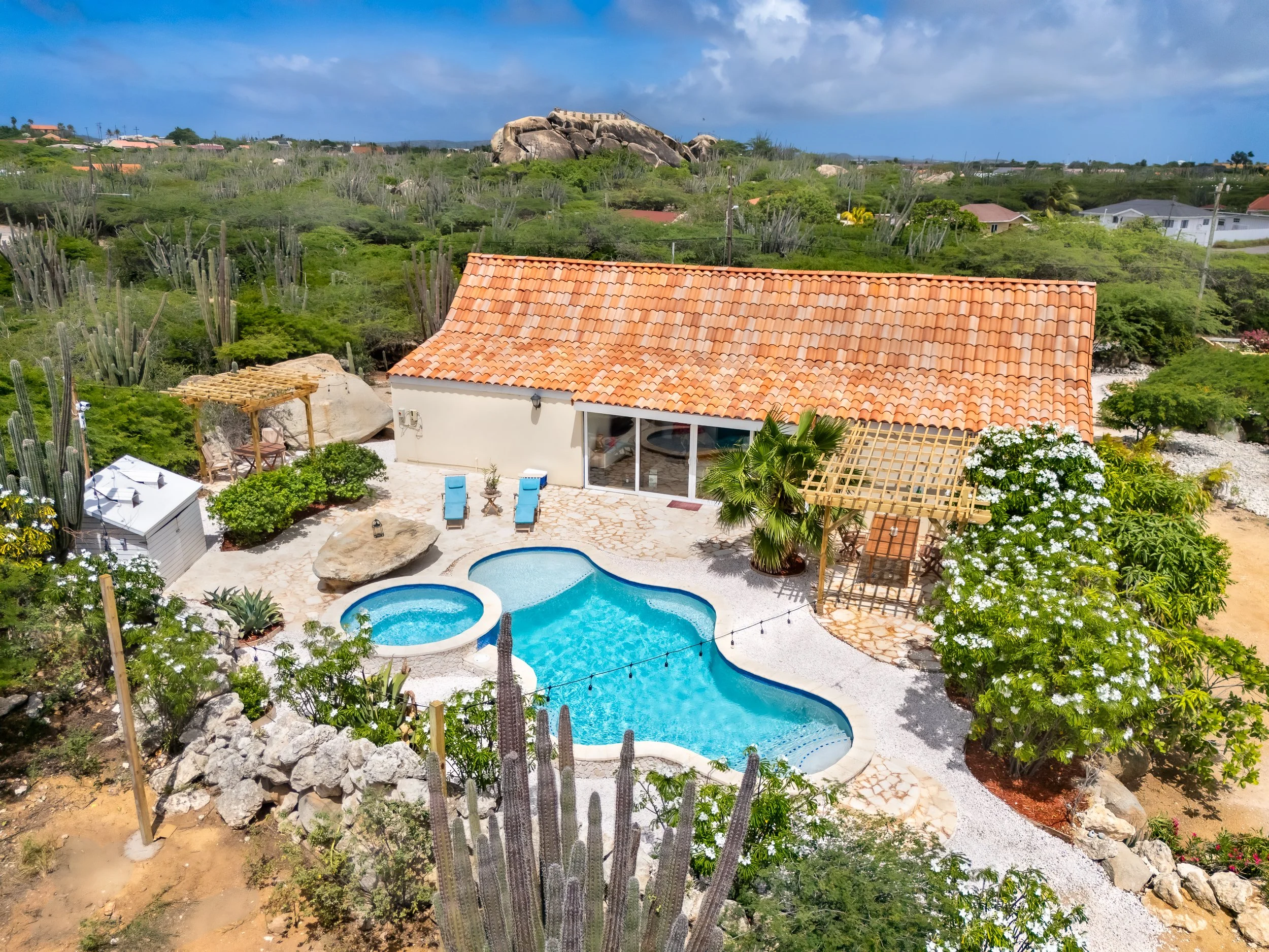 A house with a red tile roof, a backyard with a swimming pool, hot tub, two lounge chairs, and surrounding desert landscaping with cacti and green plants.