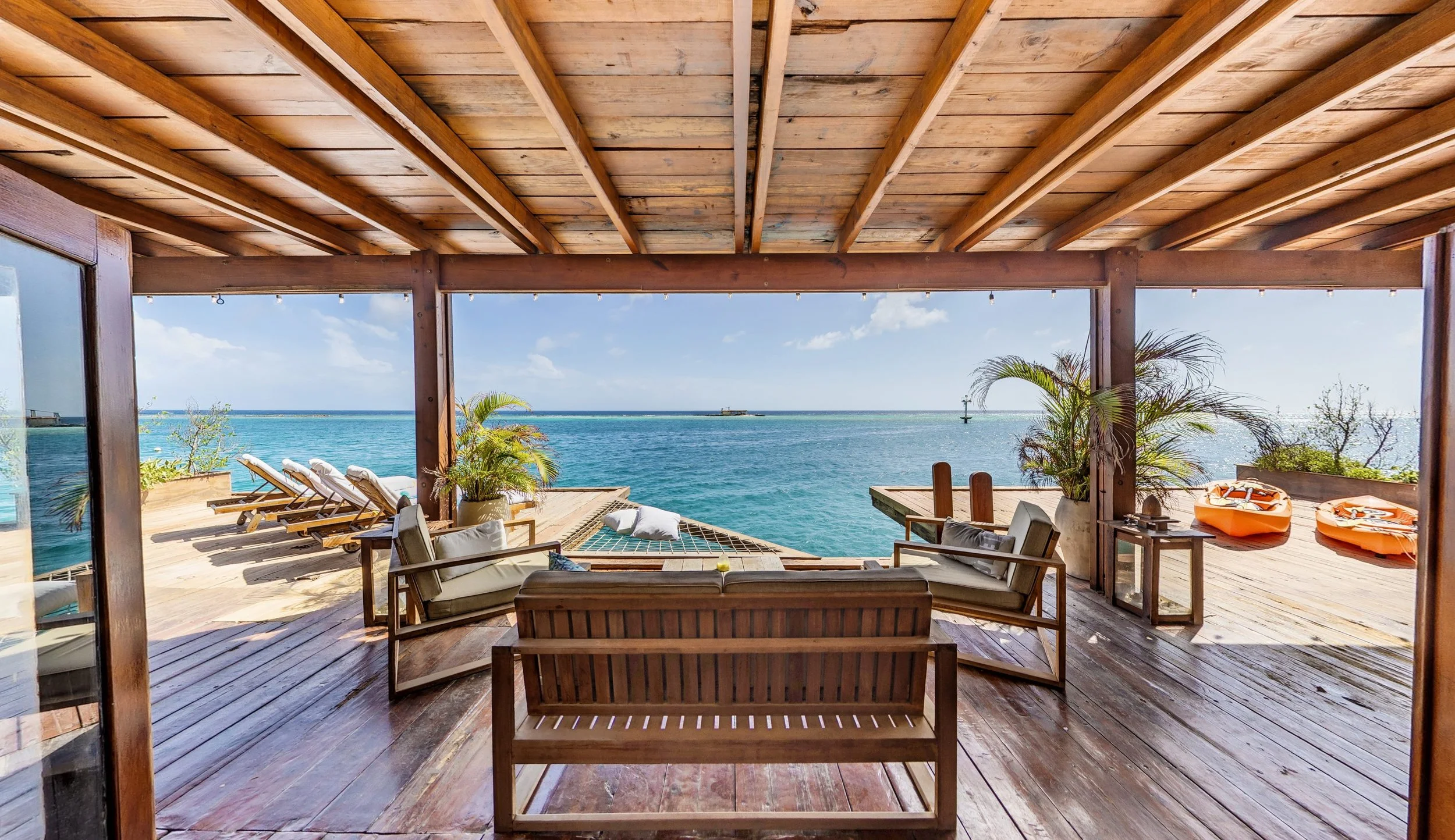 Living room with open sliding doors overlooking a sunset over the ocean, featuring cozy sofas, a wooden coffee table with plants, and tropical décor.