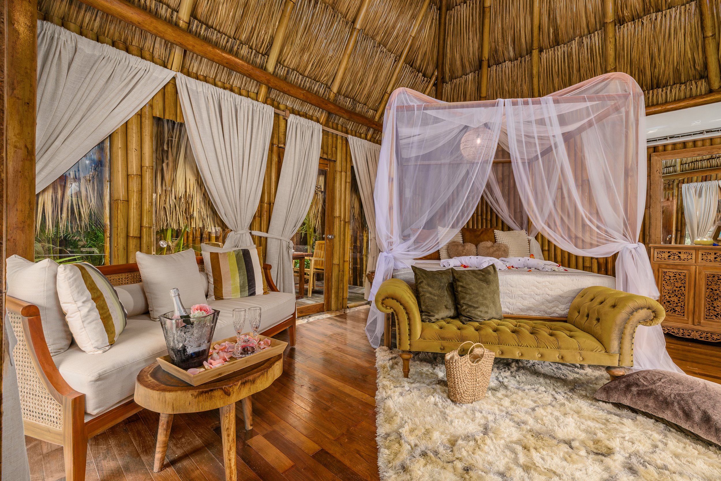 A rustic beach hut with a thatched roof, wooden walls, and a small porch with two wooden chairs with red cushions, tropical plants, and white curtains in the background.